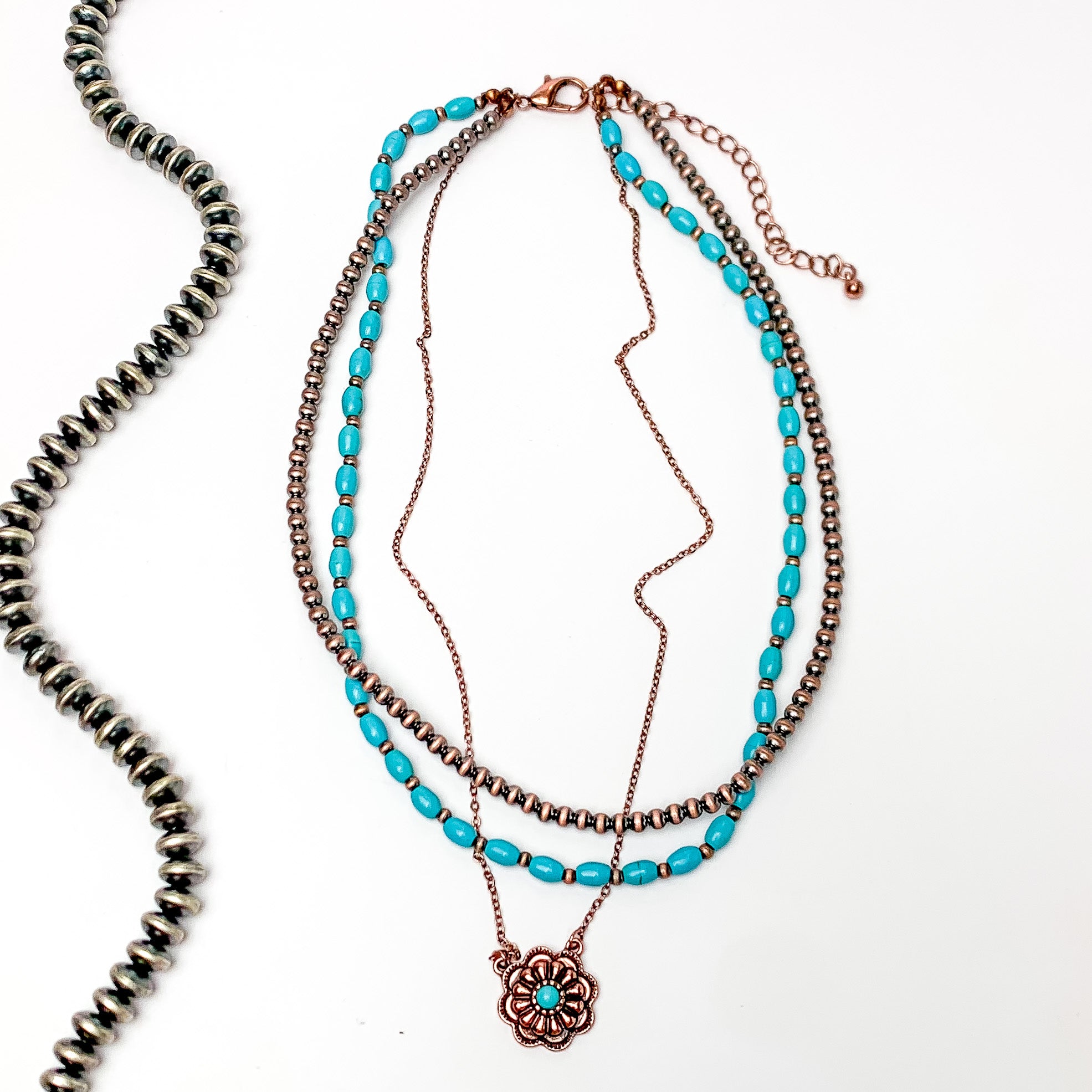 Multi strand necklace with faux navajo and turquoise beads and a dangling copper flower pendent with a turquoise stone in the middle. Pictured on a white background with a strand of copper beads to the left of it.