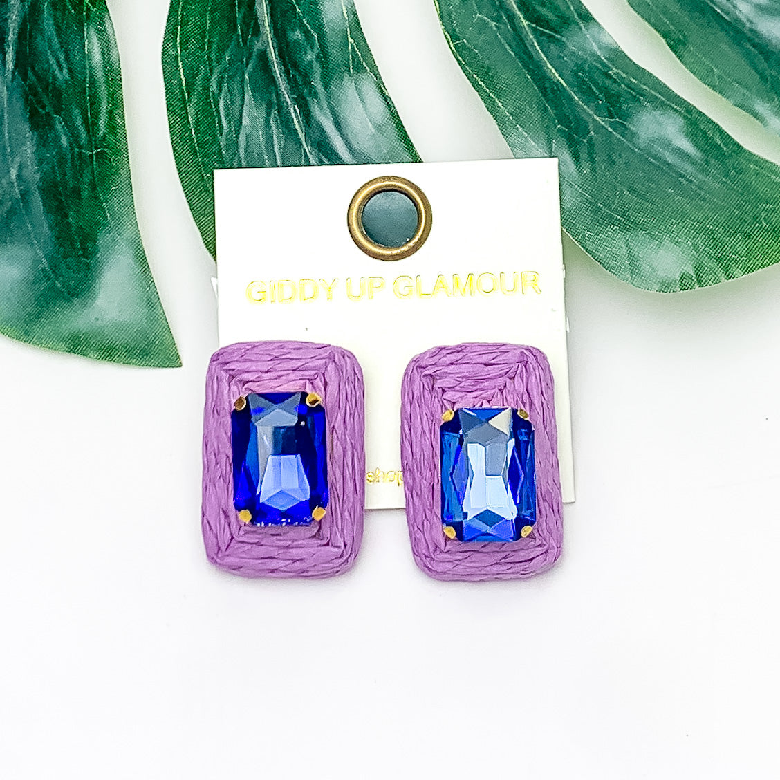 Truly Tropical Raffia Rectangle Earrings in Purple With Blue Stone. Pictured on a white background with the earrings laying on a large leaf.