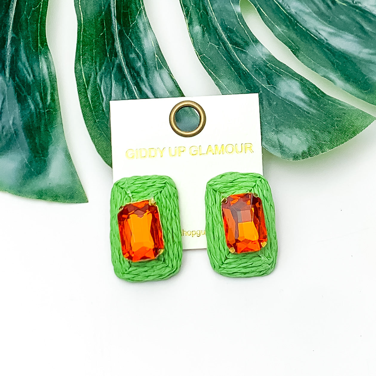 Truly Tropical Raffia Rectangle Earrings in Green With Orange Stone. Pictured on a white background with the earrings laying on a large leaf.