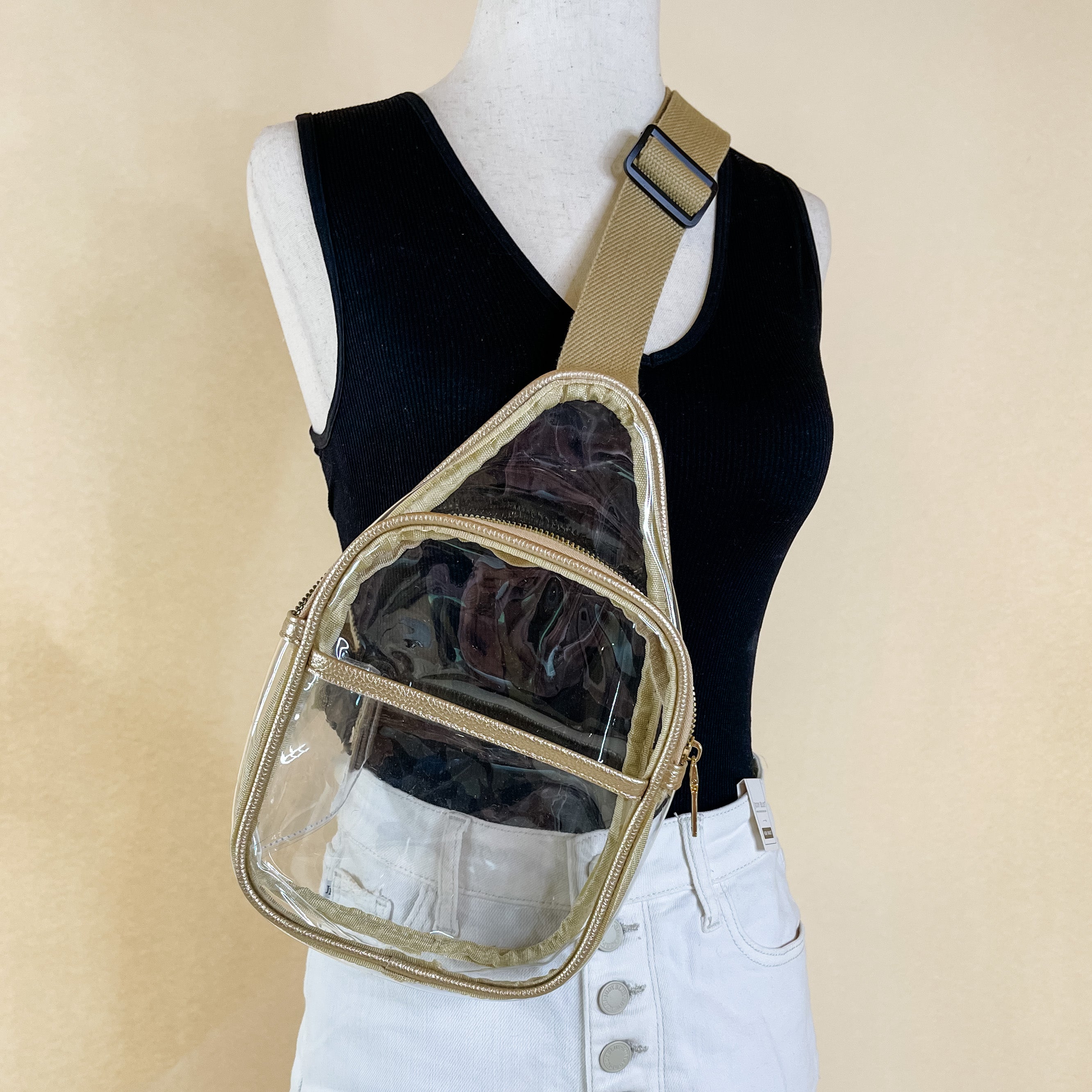Pictured is a clear sling backpack with a gold outline. This bag also includes a gold strap and black and gold accents. This bag is on a mannequin that has white shorts and a black v-neck tank pictured in front of an ivory background.  