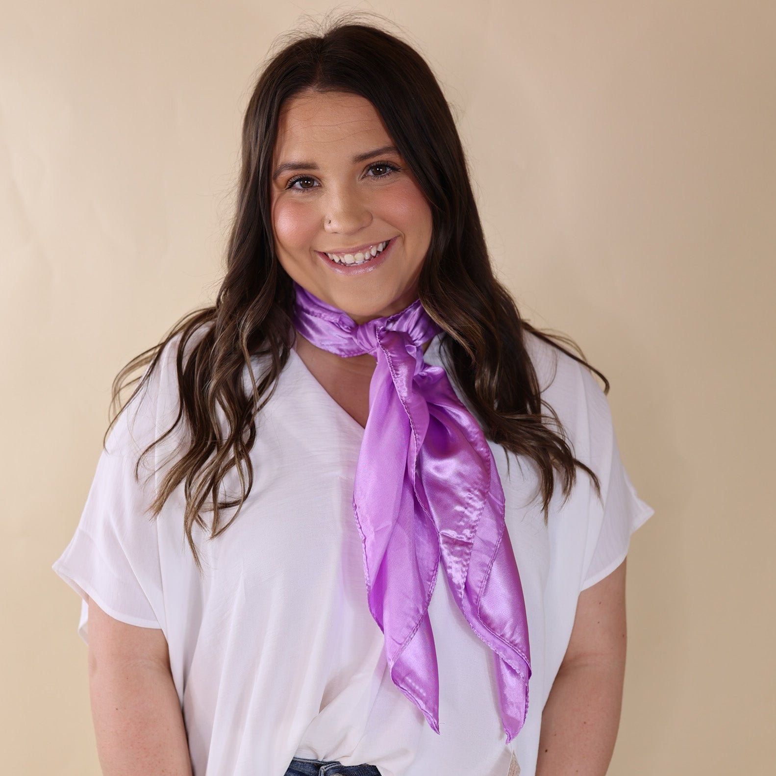 Brunette model is wearing a white drop shoulder top with a pastel purple scarf wrapped and tied around her neck. She is pictured in front of a beige background.
