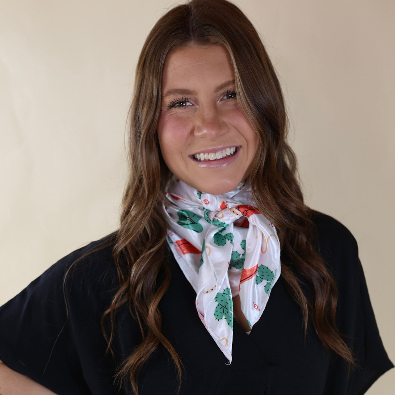 Brunette model wearing a black, drop shoulder top with a white scarf tied around her neck. The scarf includes green cacti, pink boots, and tan rope pattern. Model is pictured in front of a beige background. 