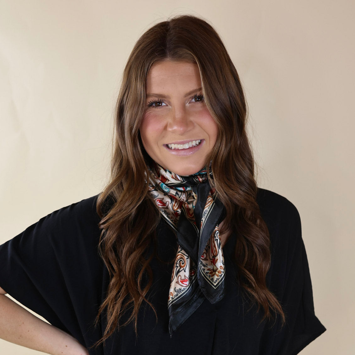 Brunette model wearing a black, drop shoulder top with beige paisley print and black outline scarf with tied around her neck. Model is pictured in front of a beige background. 