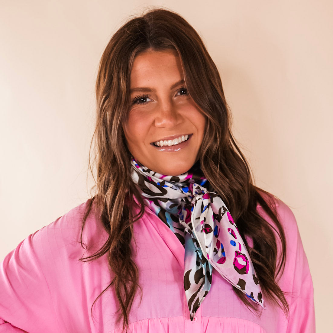Brunette model wearing a pink, button up top with a paipink and blue multicolored print scarf tied around her neck. Model is pictured in front of a beige background. 