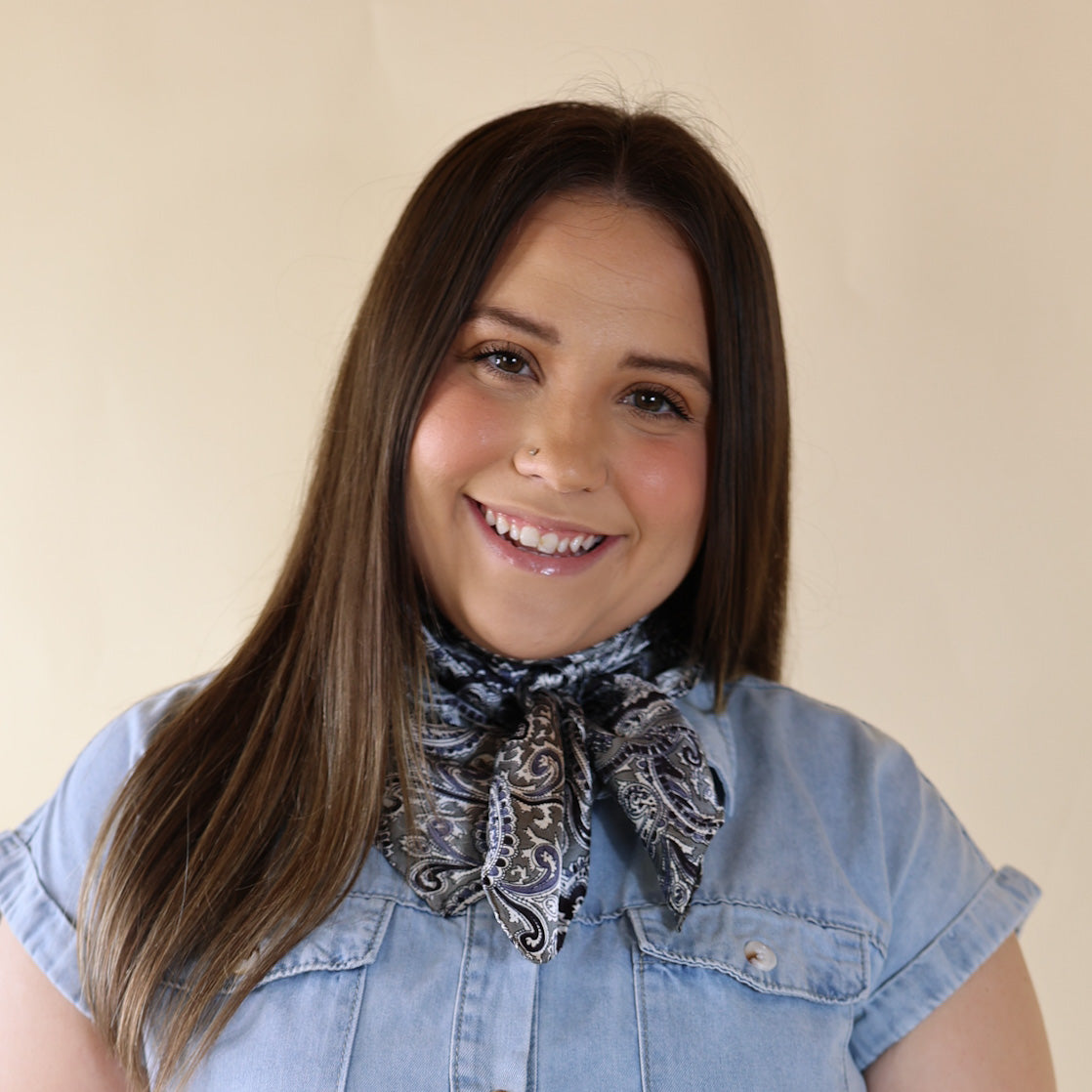 Brunette model wearing a short sleeve, denim button up with a black and silver paisly print scarf tied around her neck. This model is pictured in front of a beige background. 