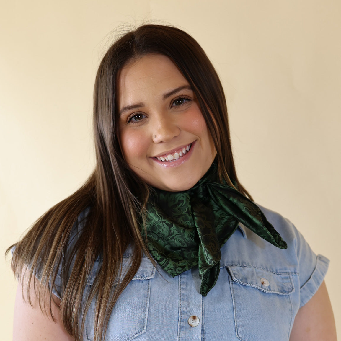 Brunette model is pictured wearing a denim button up top and a fern green, baroque printed scarf tied around her neck. She is pictured in front of a beige background. 