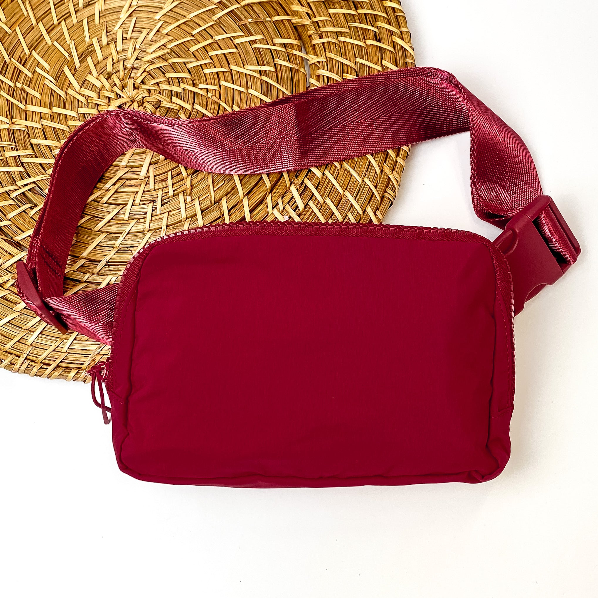 Pictured is a rectangle fanny pack with a top zipper with tassel in maroon. This bag also includes a maroon strap and maroon accents. This bag is pictured on a white and brown patterned background. 