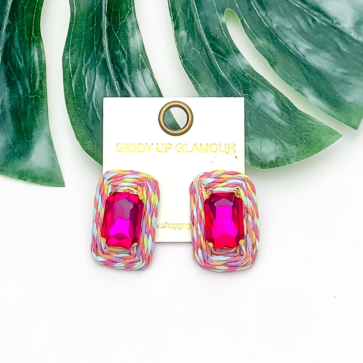 Truly Tropical Raffia Rectangle Earrings in Multicolor With Fuchsia Stone. Pictured on a white background with the earrings laying on a large leaf.