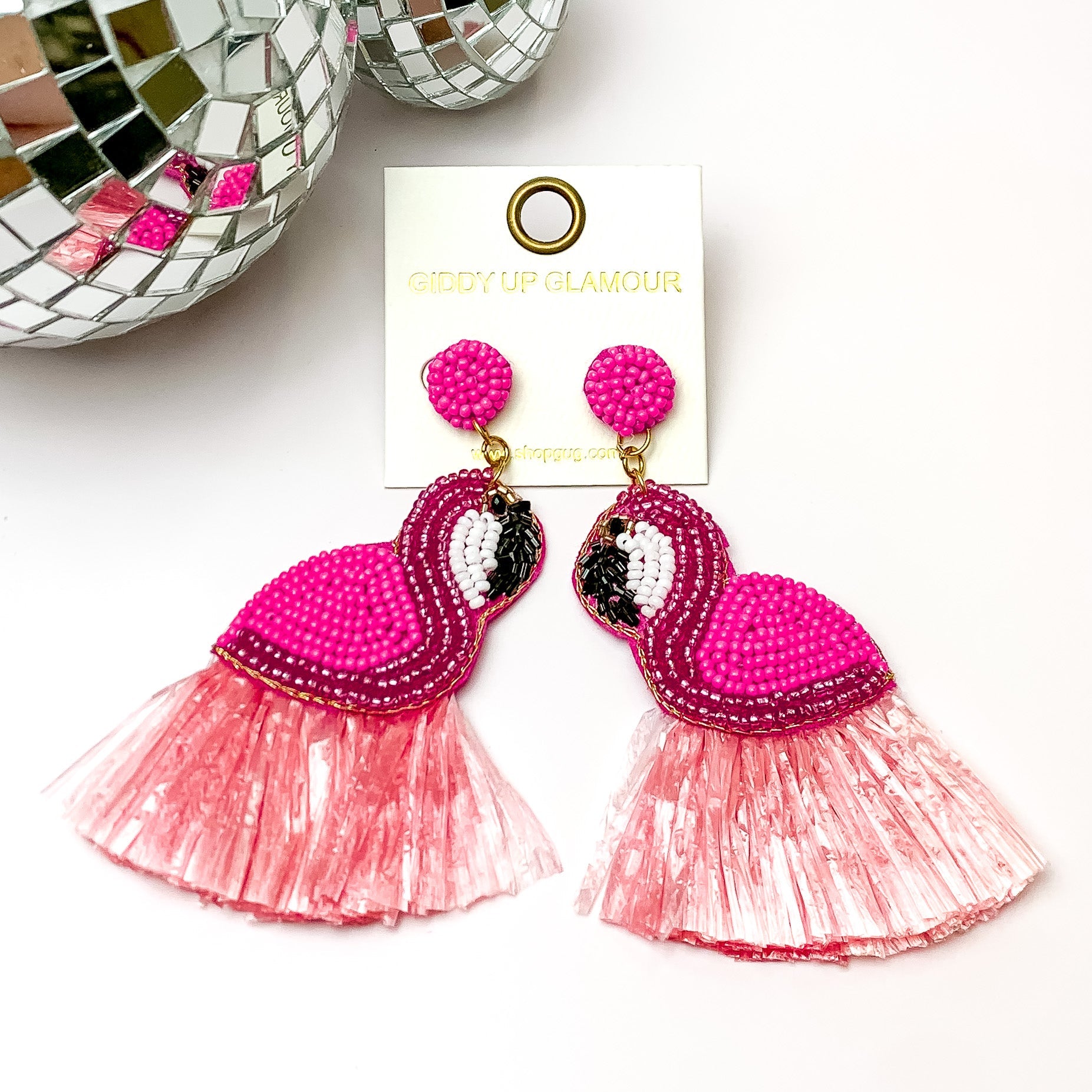 Statement pink flamingo earrings with pink fringe. Pictured on a white background with a disco ball at the top left.