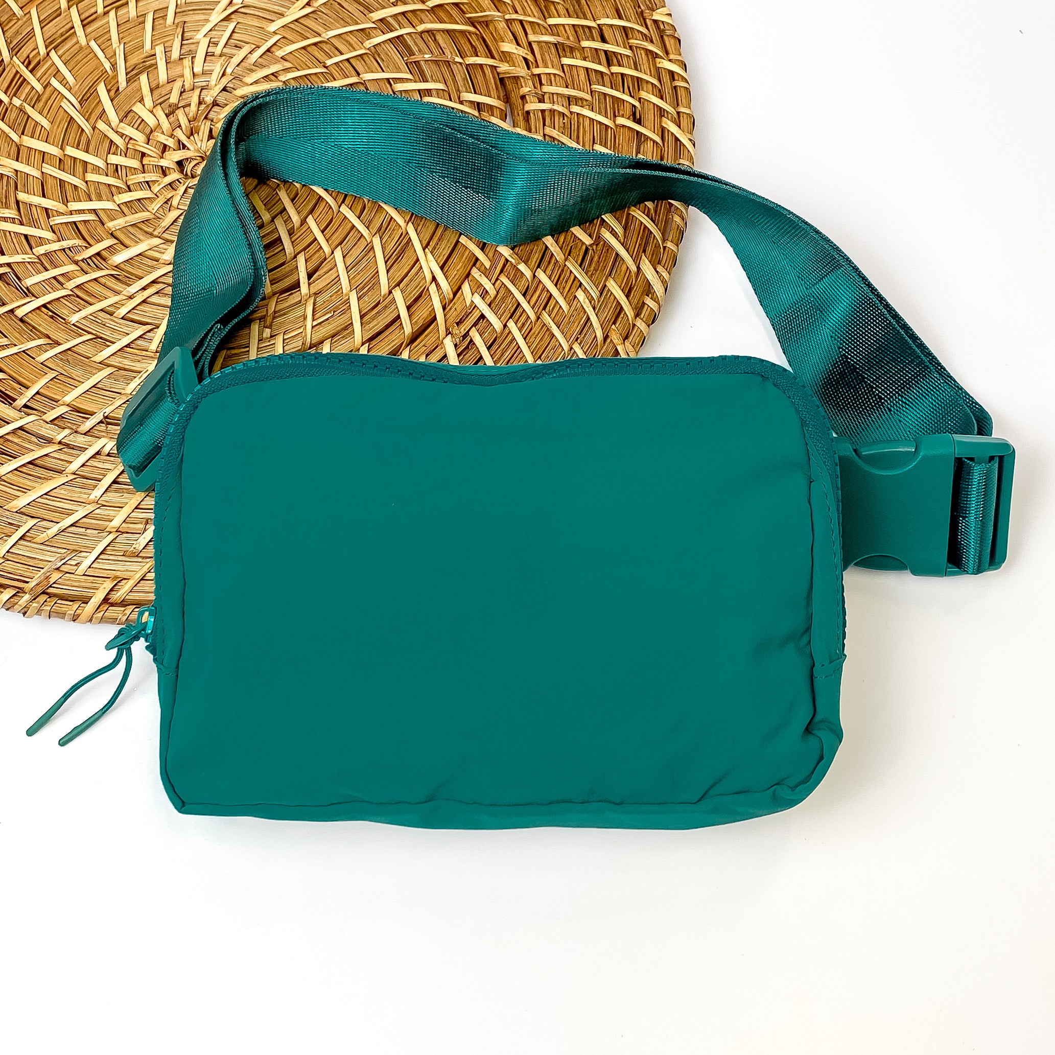 Pictured is a rectangle fanny pack with a top zipper with tassel in everglade green. This bag also includes a everglade green strap and everglade green accents. This bag is pictured on a white and brown patterned background. 
