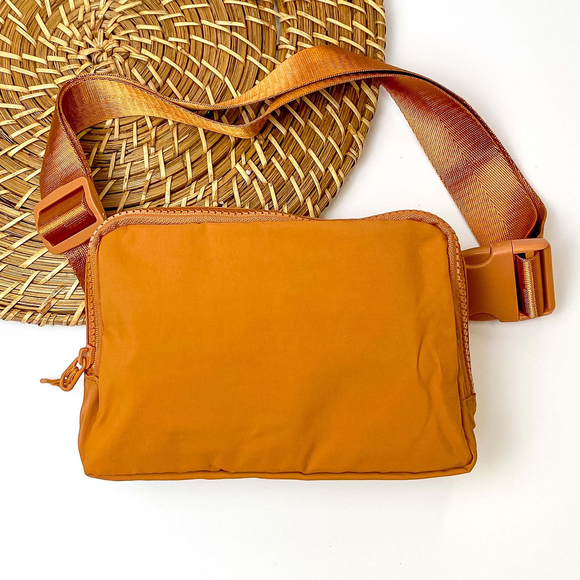 Pictured is a rectangle fanny pack with a top zipper with tassel in tan. This bag also includes a tan strap and tan accents. This bag is pictured on a white and brown patterned background. 