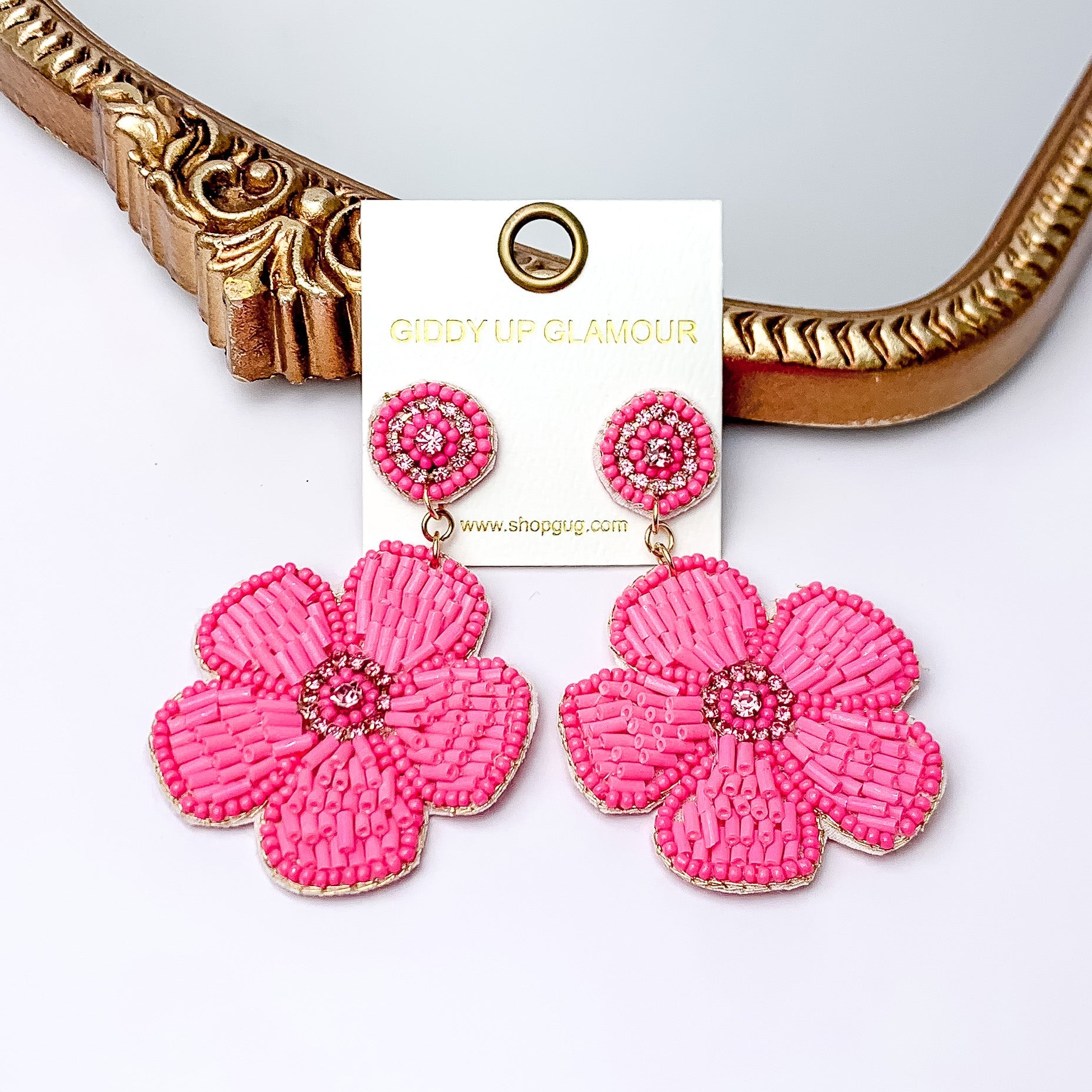 There is hanging beaded flower pendant from the gold stud earrings. The flower pendants are pink with  gold outline and detailing. These earrings are pictured in front of a gold mirror on a white background.