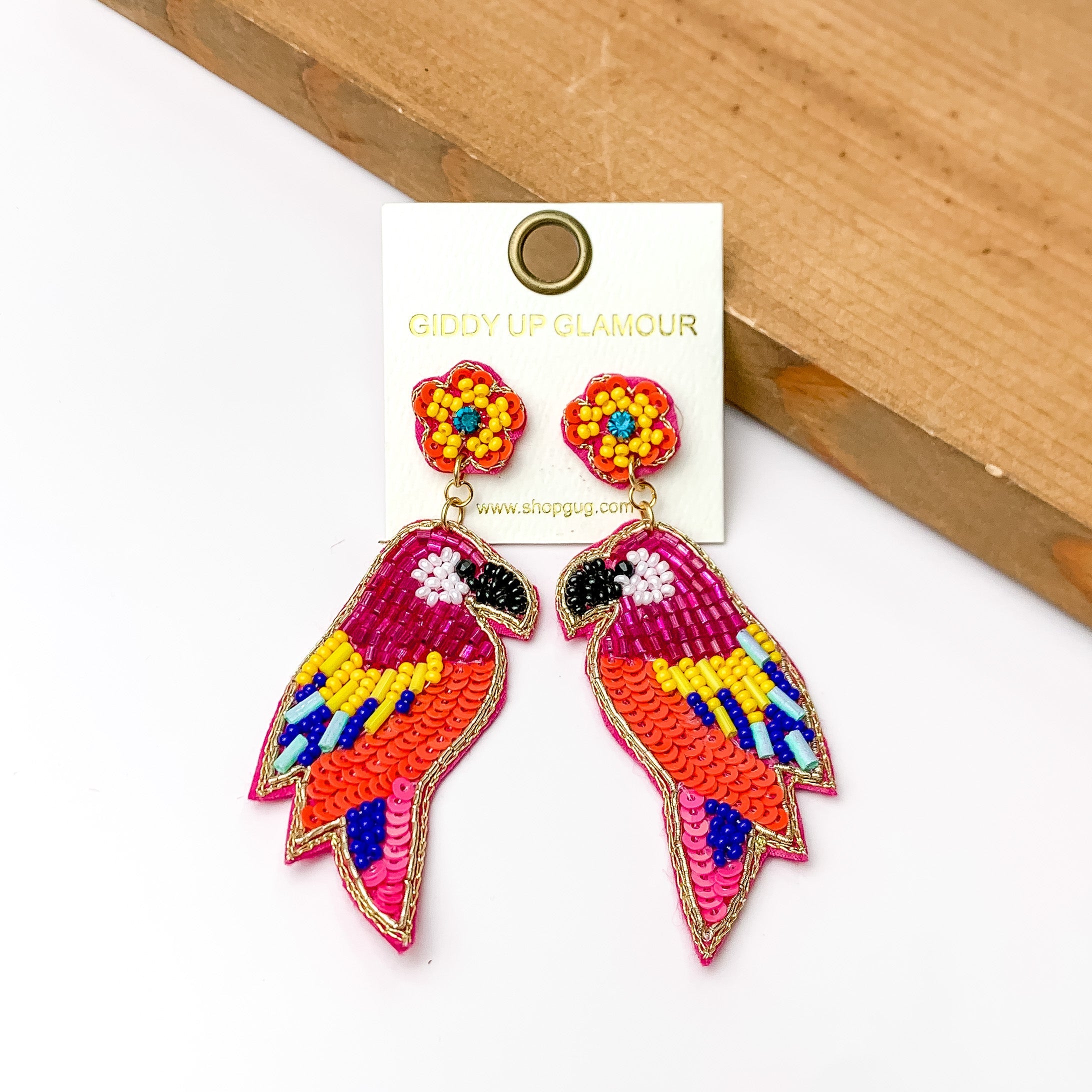 These parrot earrings are pink with beads that are yellow, turquoise, and royal blue. They do have seqence that are pink and orange. These earrings are pictured in front of a piece of wood with a white background.
