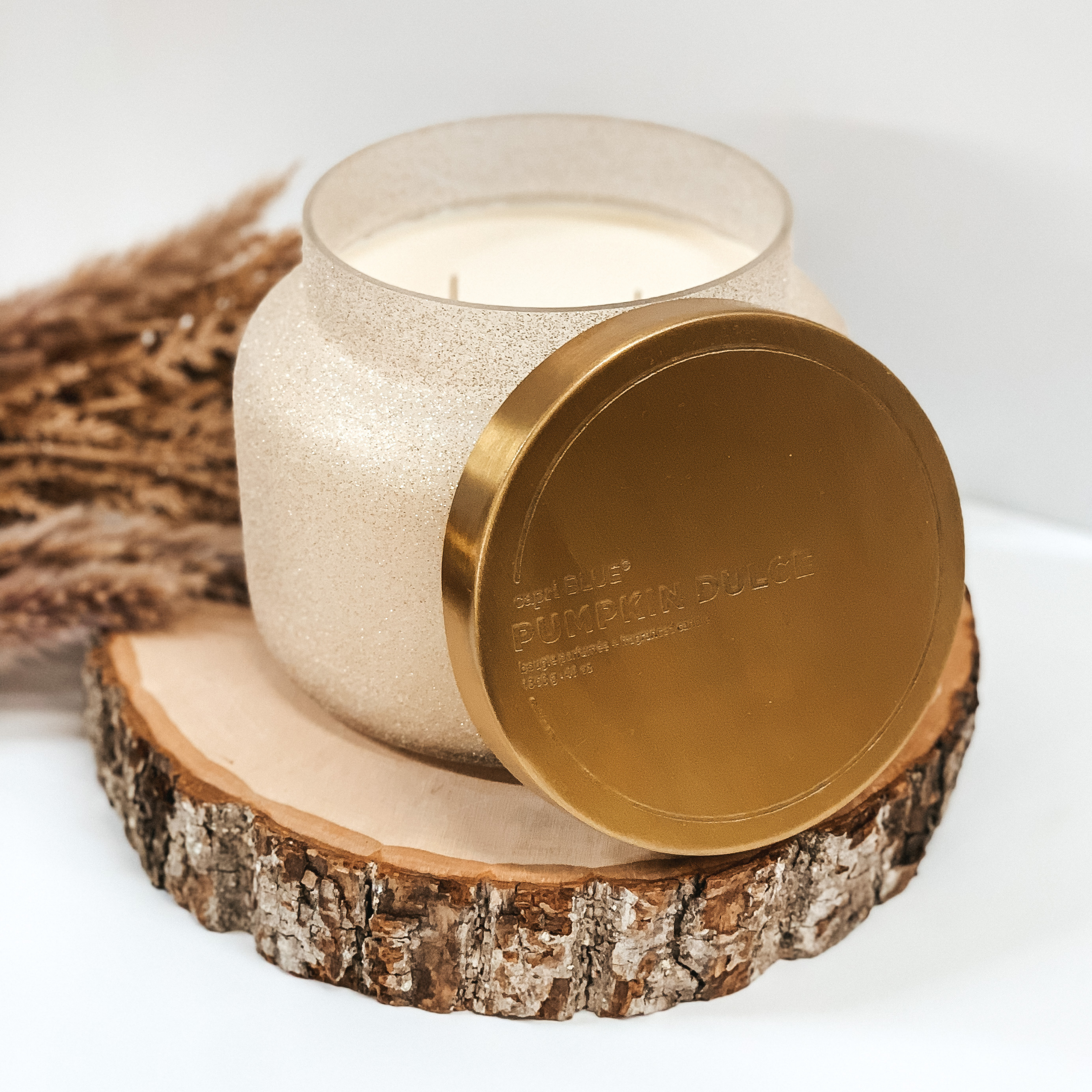 Champagne glitter candle with a gold lid pictured on a white background on a piece of wood. 