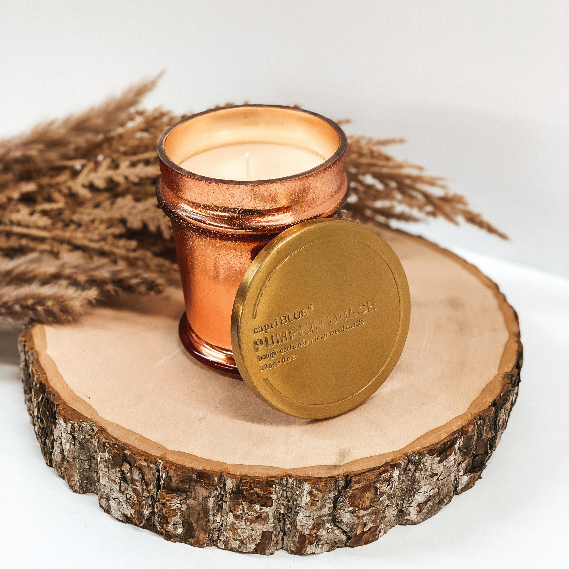 Copper glitter candle and a gold lid pictured on a white background on a piece of wood with brown pompous grass in behind the candle. 