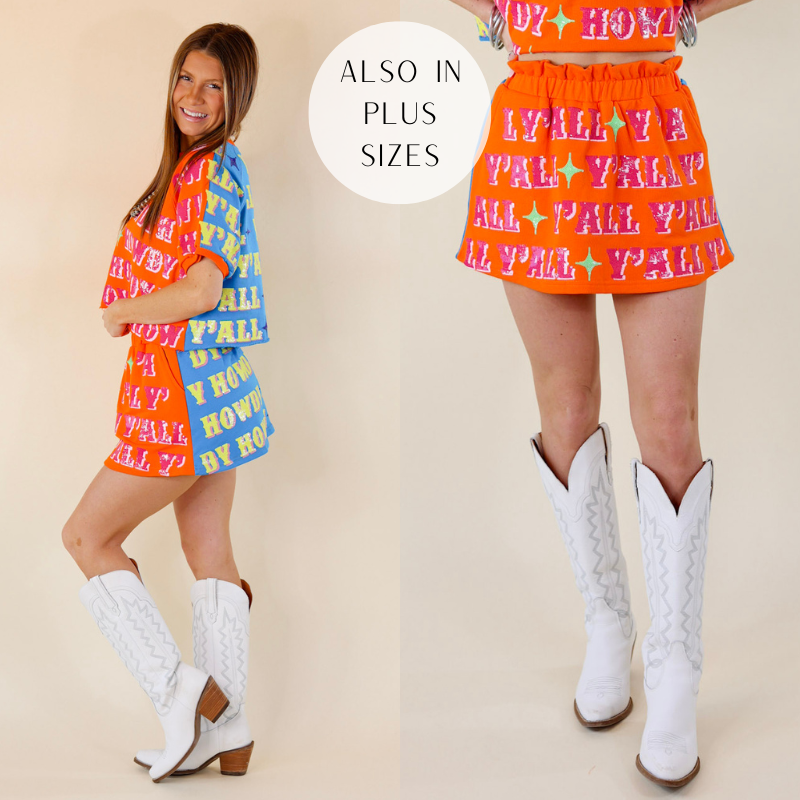 Model is wearing a colorblock skort with the word "Howdy" and "Yall" in pink and yellow on orange and blue fabric. Model has it paired with white boots, matching top, and silver jewelry.
