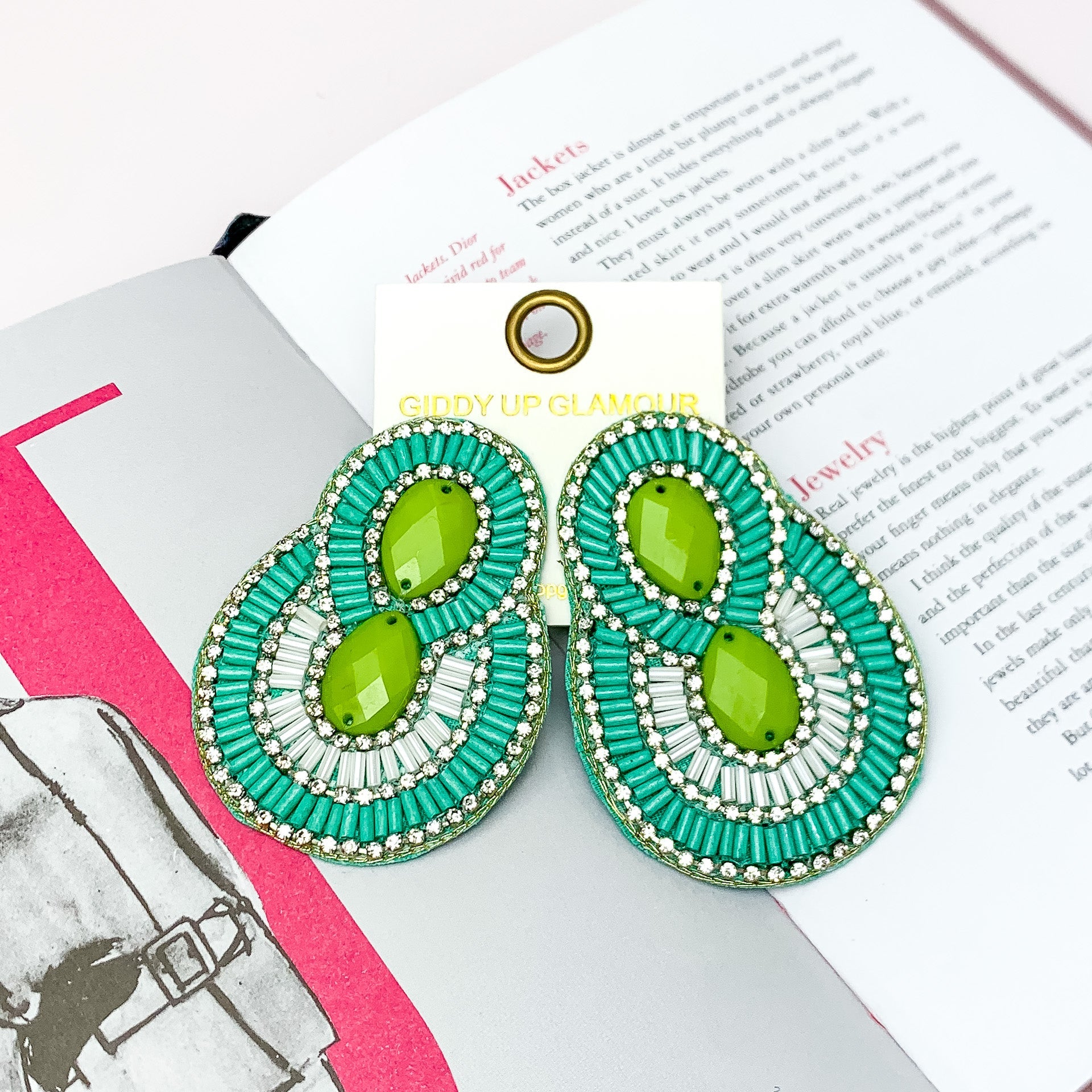 Dazzling Oval Earrings Outlined in Clear Crystals with Beads in Green. Pictured on a white background with a book behind the earrings. 