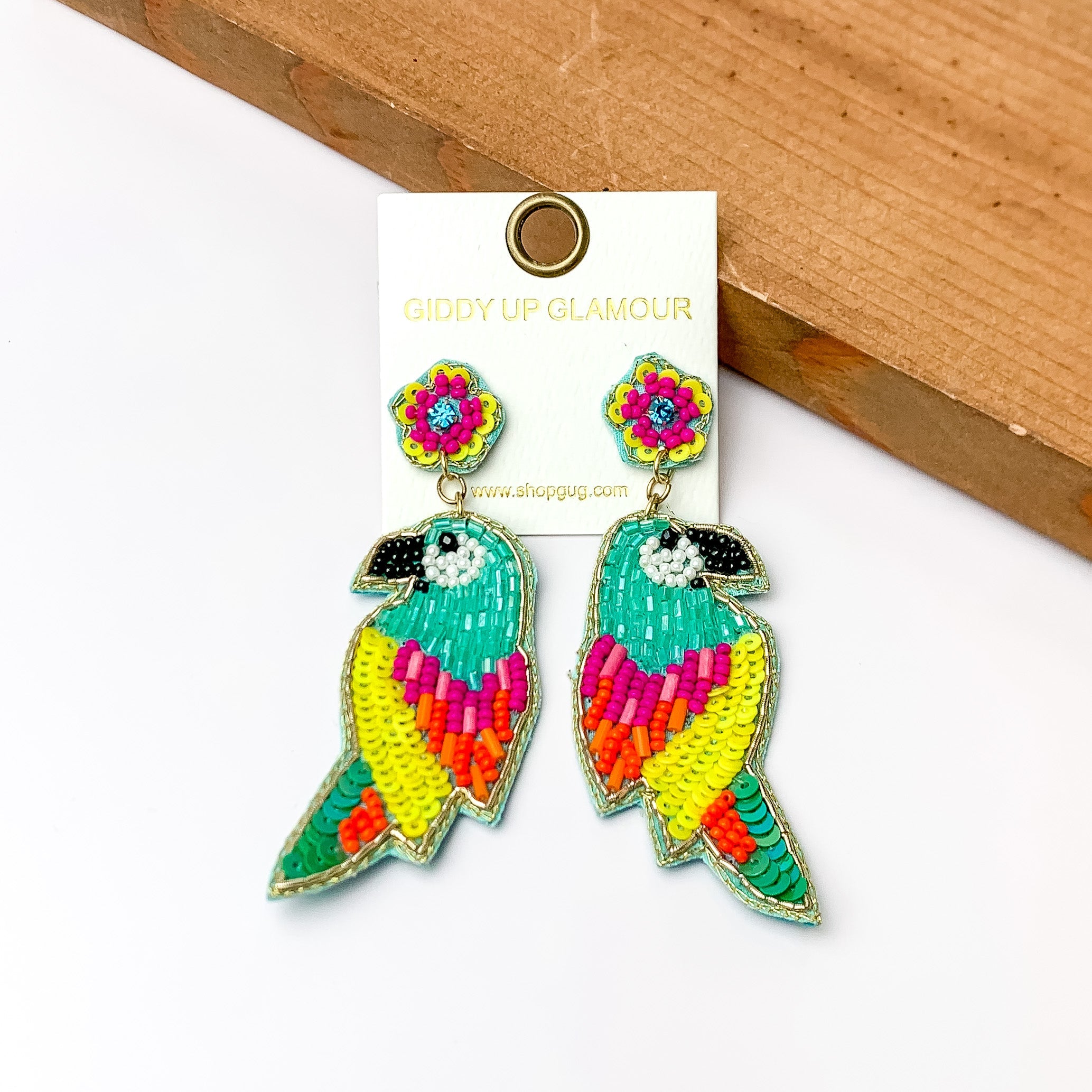 These parrot earrings are turquoise with beads that are yellow, fush, and orange. They do have seqence that are neon yellow and dark turquoise. These earrings are pictured in front of a piece of wood with a white background.
