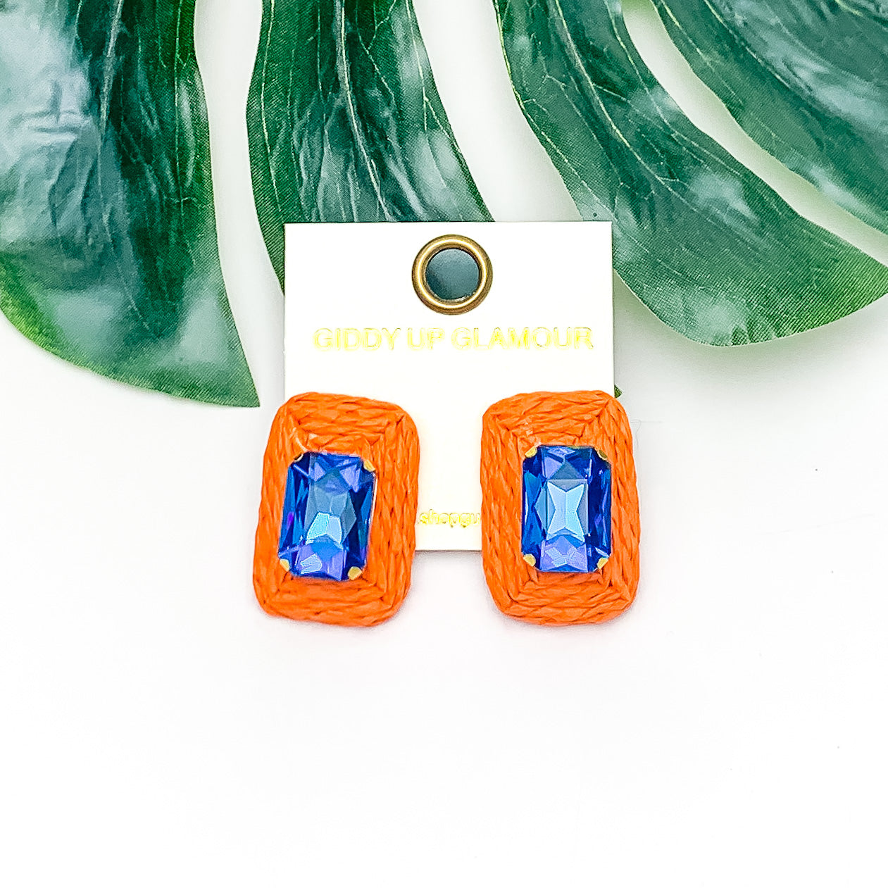 Truly Tropical Raffia Rectangle Earrings in Orange With Blue Crystal. Pictured on a white background with the earrings laying on a large leaf.