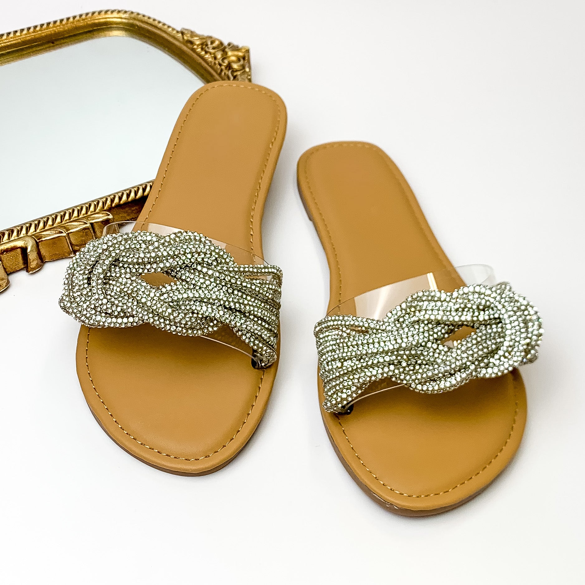 Tan sandals with a clear band that has clear crystals bands tied in a knot on top of the clear band. These sandals are pictured on a white background with a gold mirror in the top left corner. 