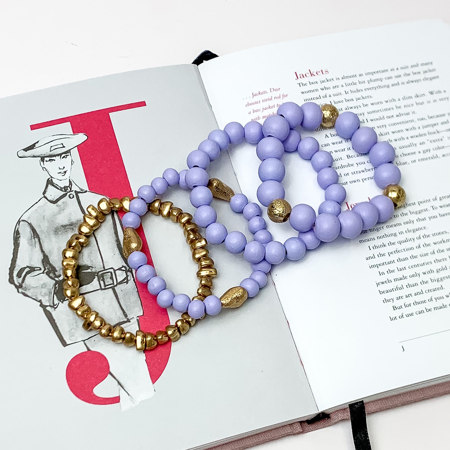 Set of Four | Stretchy Light Purple Beaded Bracelets featuring a Gold Tone Bracelet. Pictured on a white background. Bracelets are laying on top of an open book.