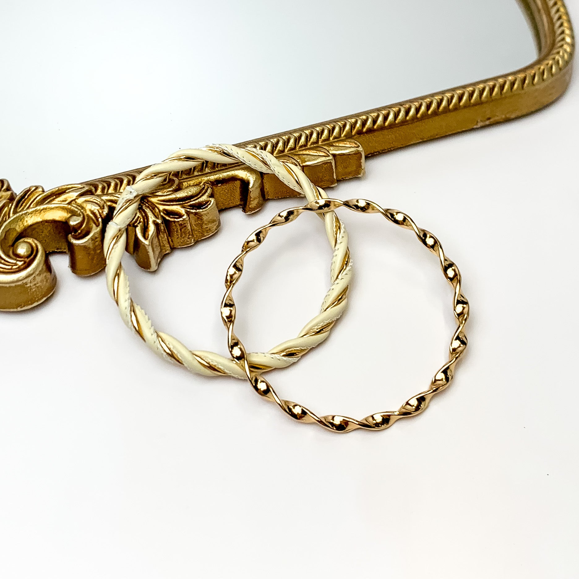 Pictured is a gold twist bangle and gold and ivory twist bangle. These bangles are pictured partially leaning on a gold mirror on a white background. 