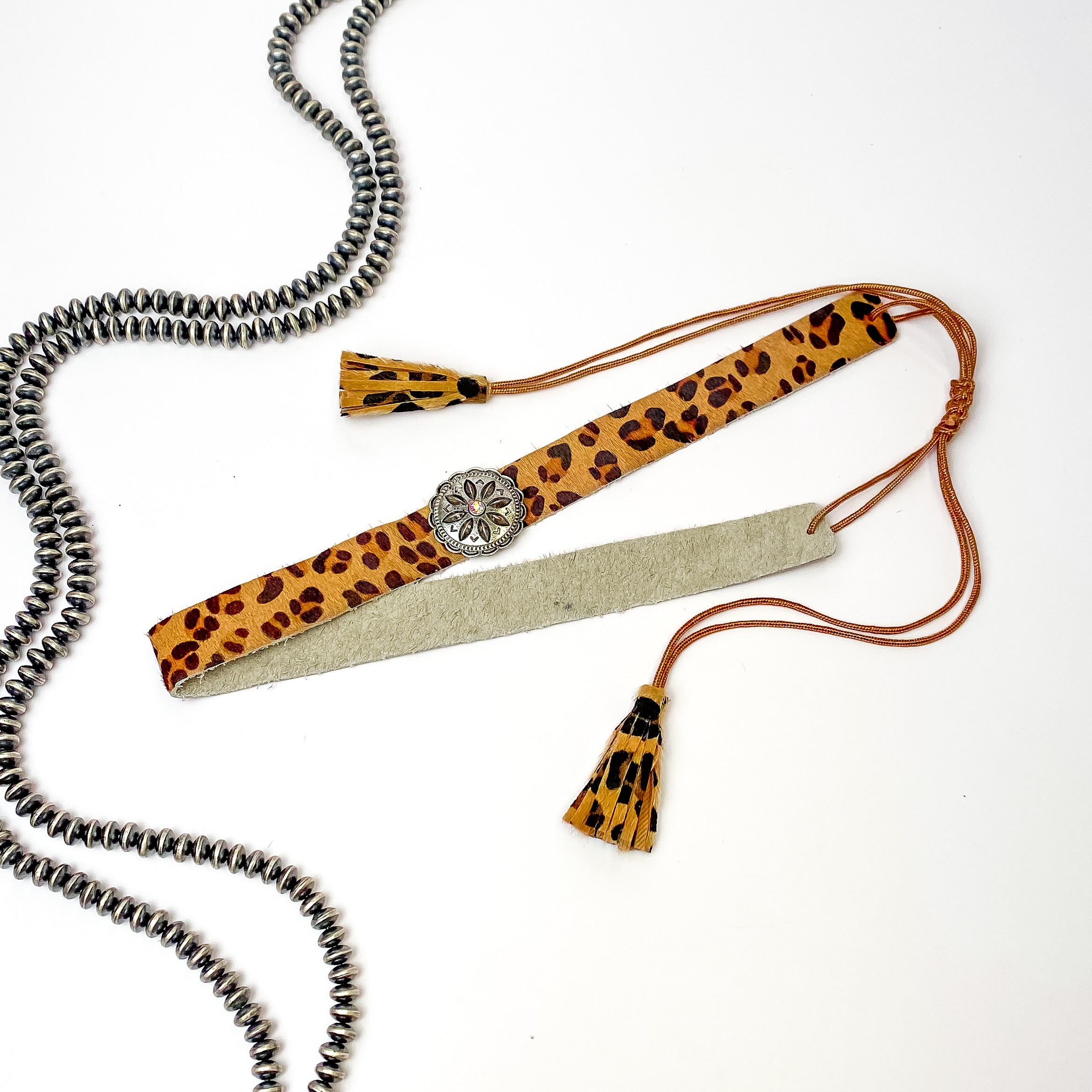 Pictured is a leopard print, adjustable hat band with a silver concho. This hat band is pictured on a white background with silver, saucer beads on the left side of the hat band. 