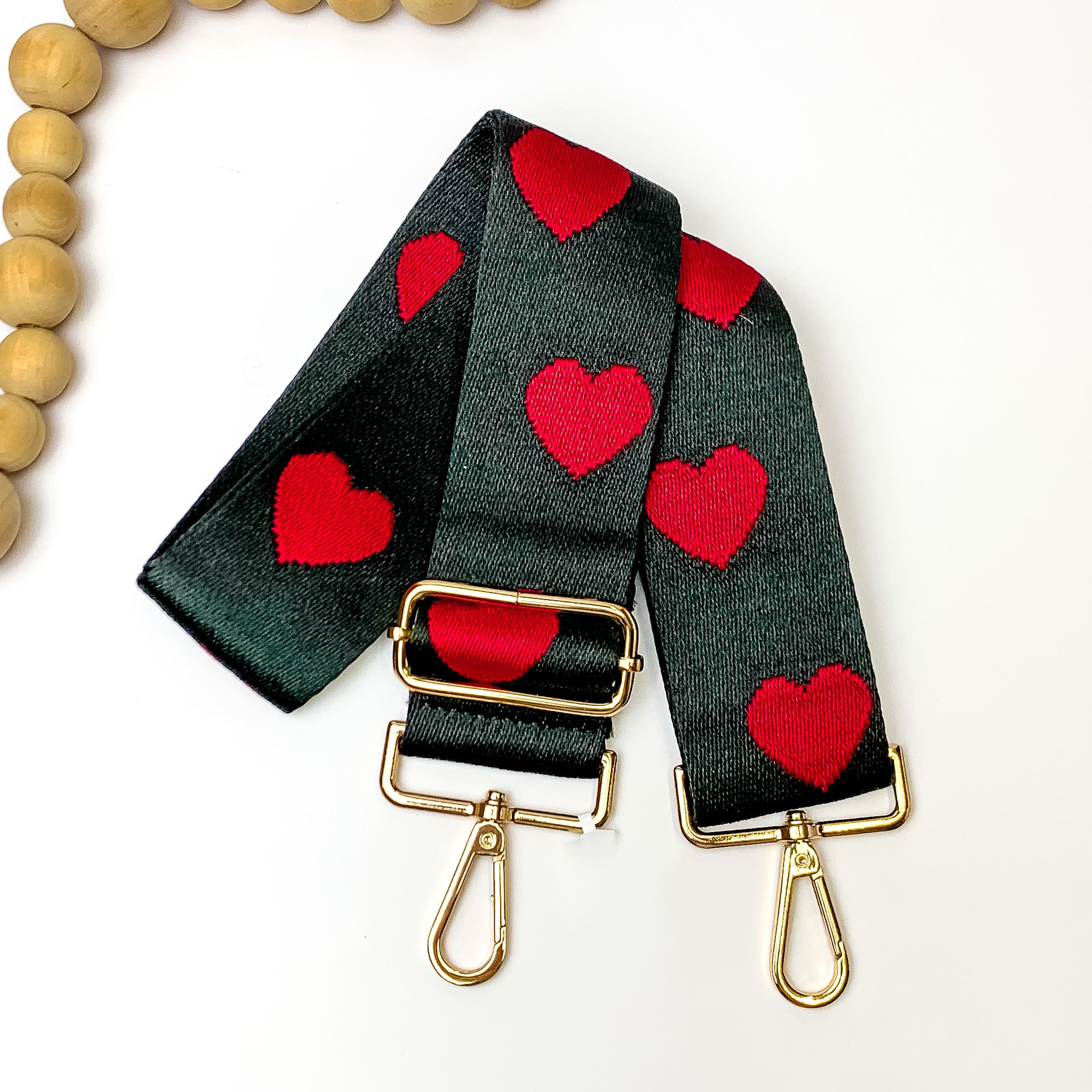 Black purse strap with a red heart design. This purse strap includes gold hardware. This purse strap os pictured on a white background with tan beads in the top left corner. 