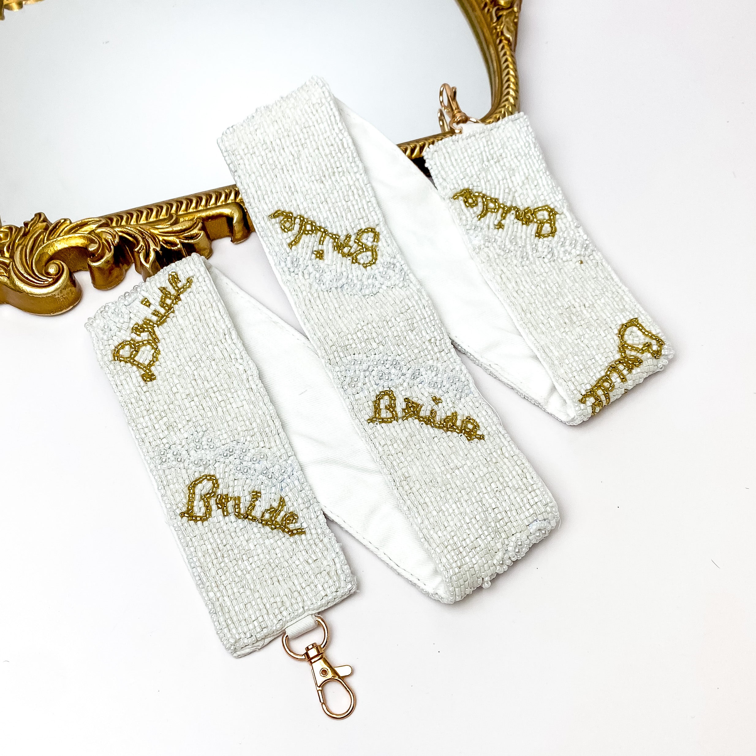White beaded purse strap with gold hardware and the word "Bride" in gold beads. This purse strap is pictured partially laying on a gold mirror on a white background. 