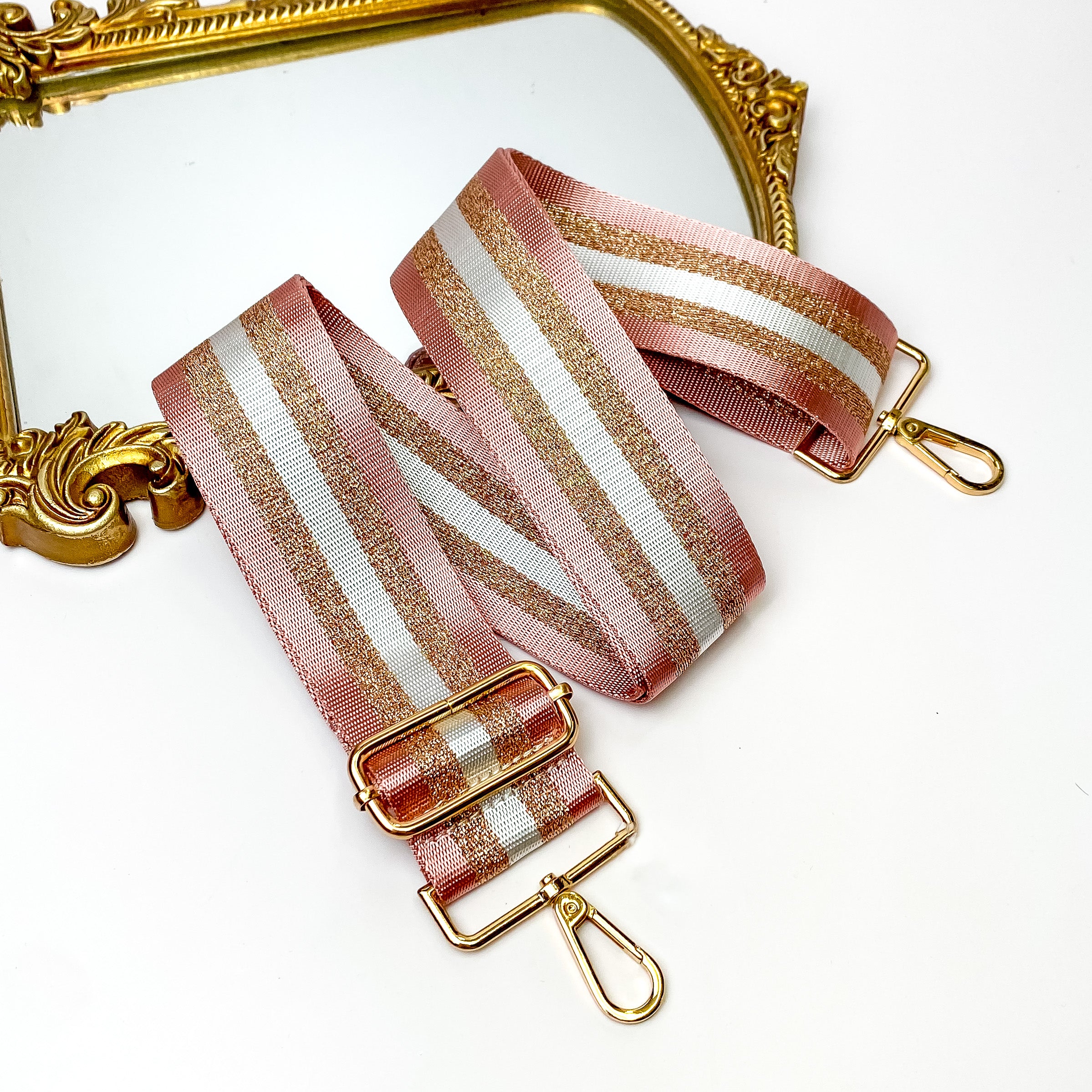 Pictured is a striped purse strap with gold accessories. This purse strap has two outer pink stripes, then two gold glitter stripes, and a center white stripe. This purse strap is laid out partially on a gold mirror on a white background. 