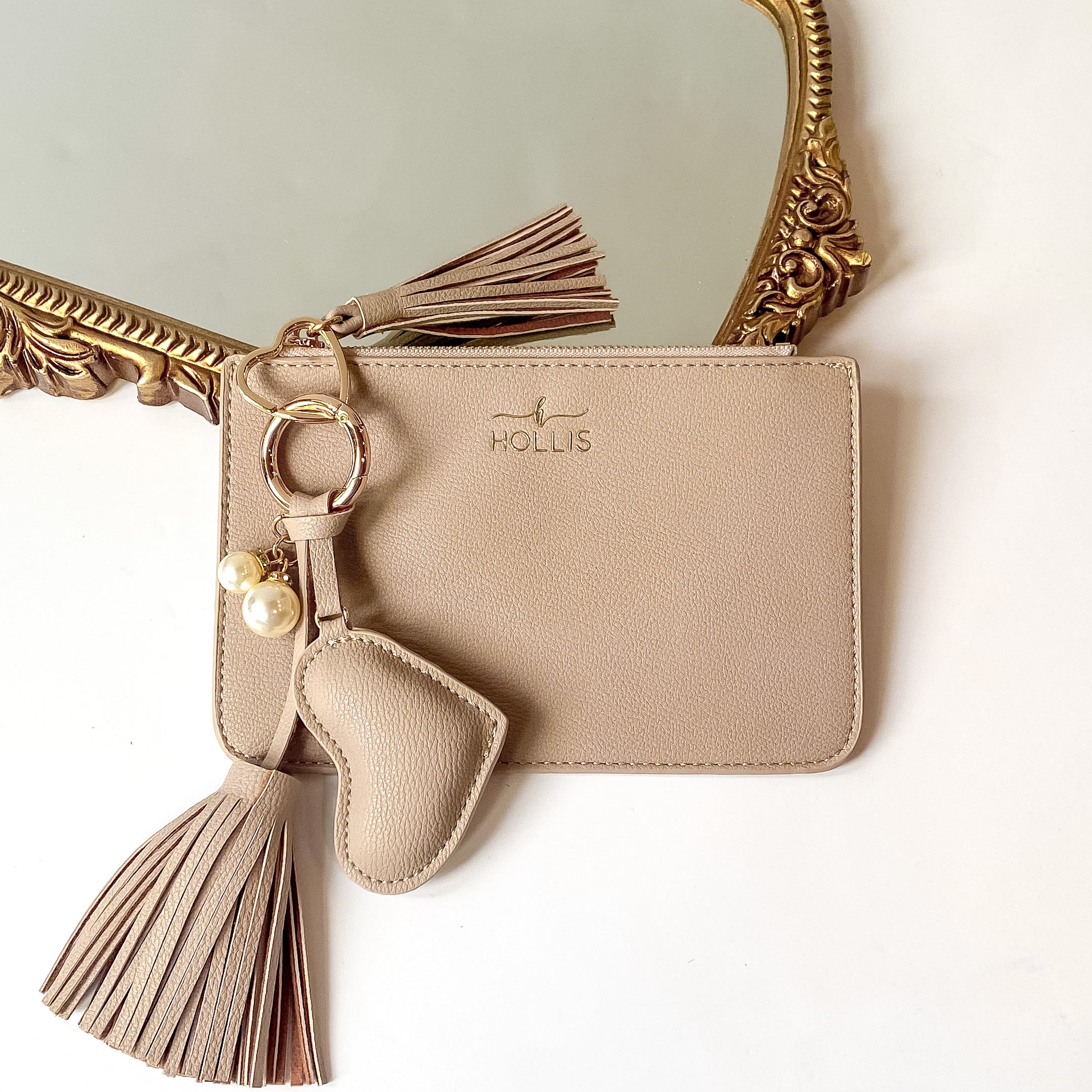 Nude coin pouch with a gold HOLLIS emblem at the top and a top zipper. This coin pouch has a gold key ring, gold heart key ring, a nude tassel charm, a nude heart charm and white pearl charms. This luggage tag is pictured partially laying on a gold mirror on a white background. 