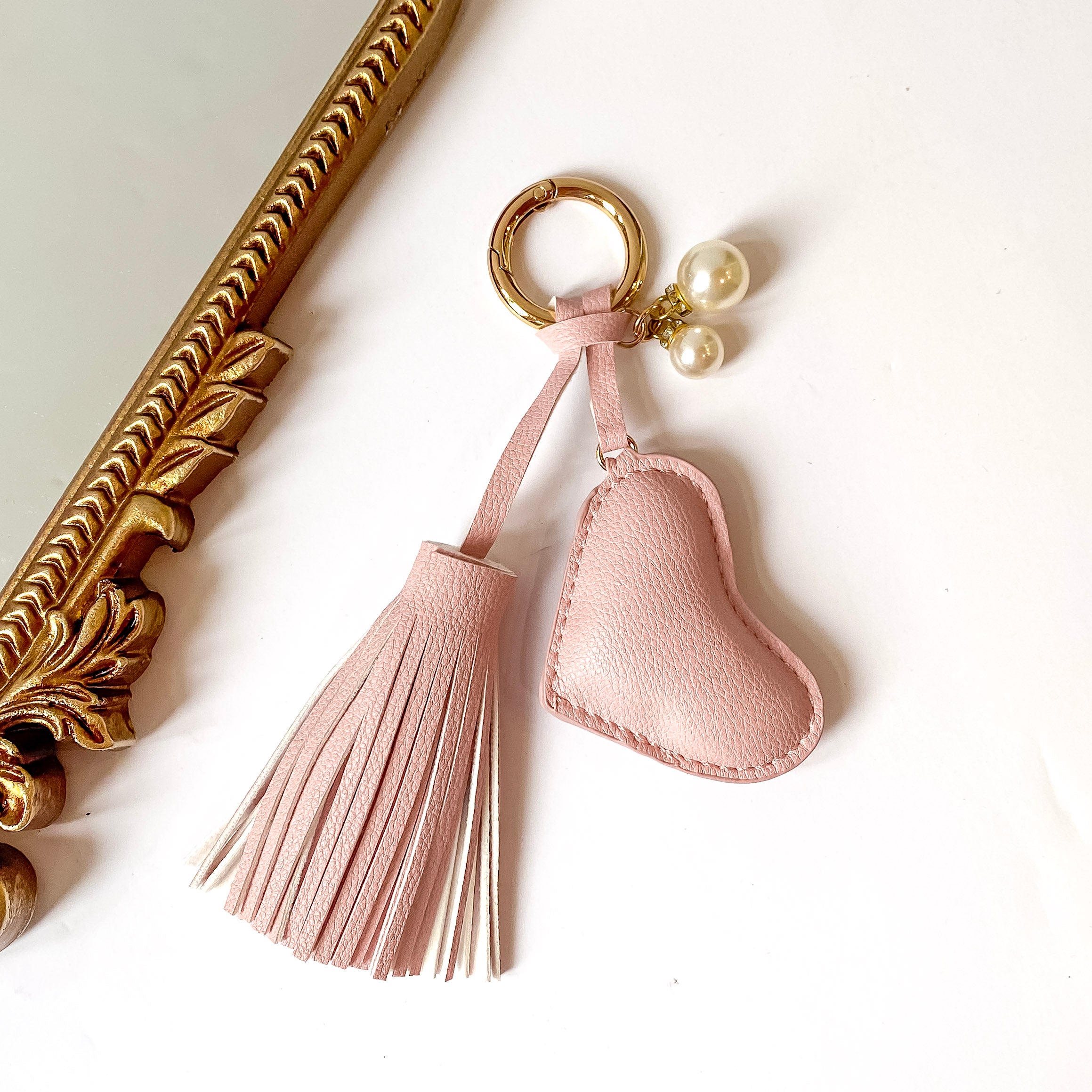 Gold key ring with a blush pink tassel and heart charm. This key ring also includes two white, pearl charms. This is pictured on a white background with a gold mirror on the left side of the picture. 