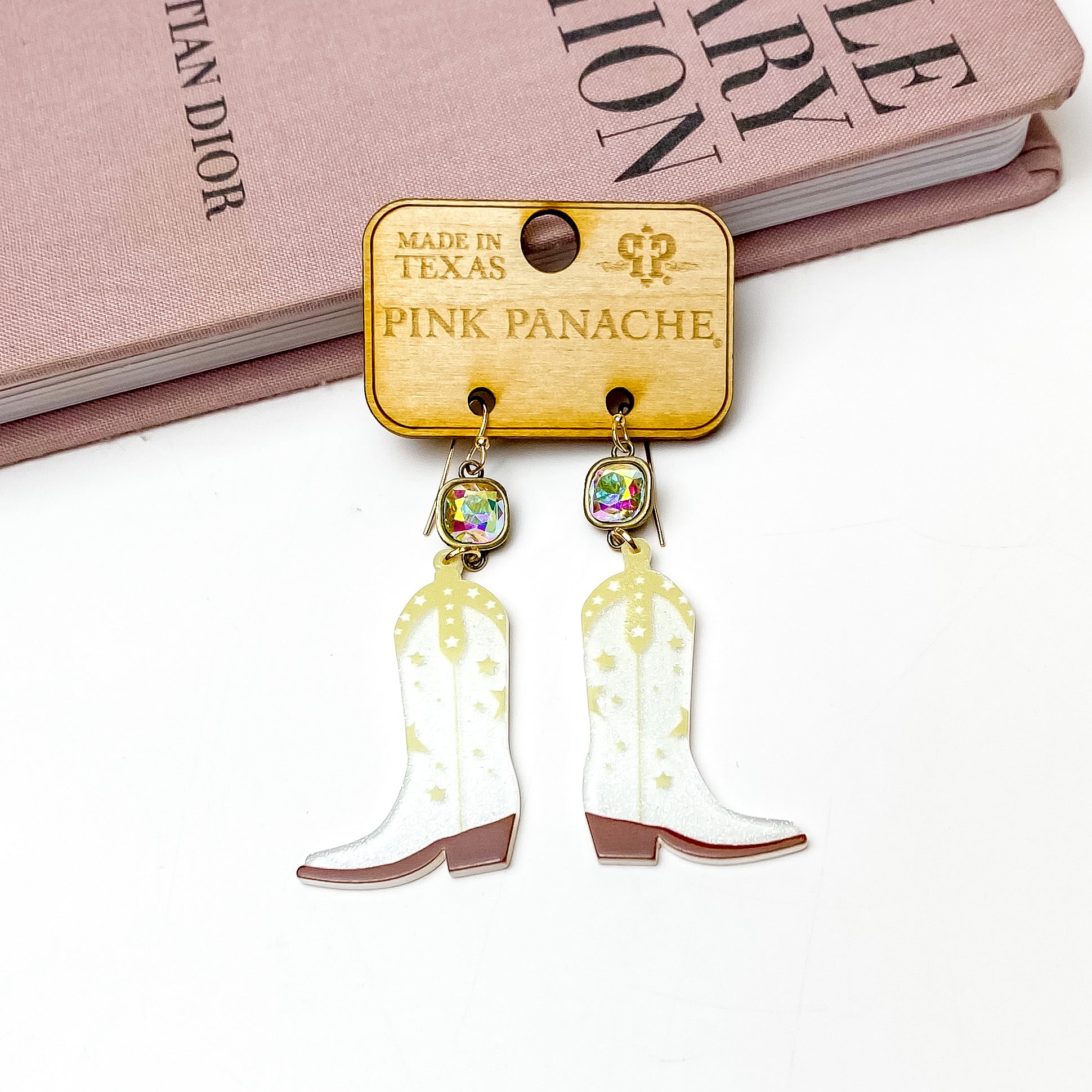 AB cushion cut crystal drop earrings with a white glitter boot pendants. These boot pendants have cream and brown accents. These earrings are pictured on a wooden earring holder on a white background in front of a mauve colored book. 