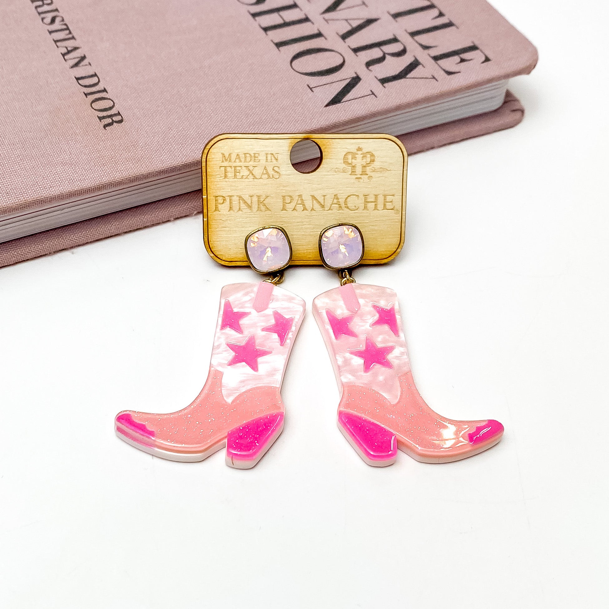 Rose water opal cushion cut crystal post earrings with a pink pearlized and glitter boot pendants. These boot pendants have pink accents. These earrings are pictured on a wooden earring holder on a white background in front of a mauve colored book. 