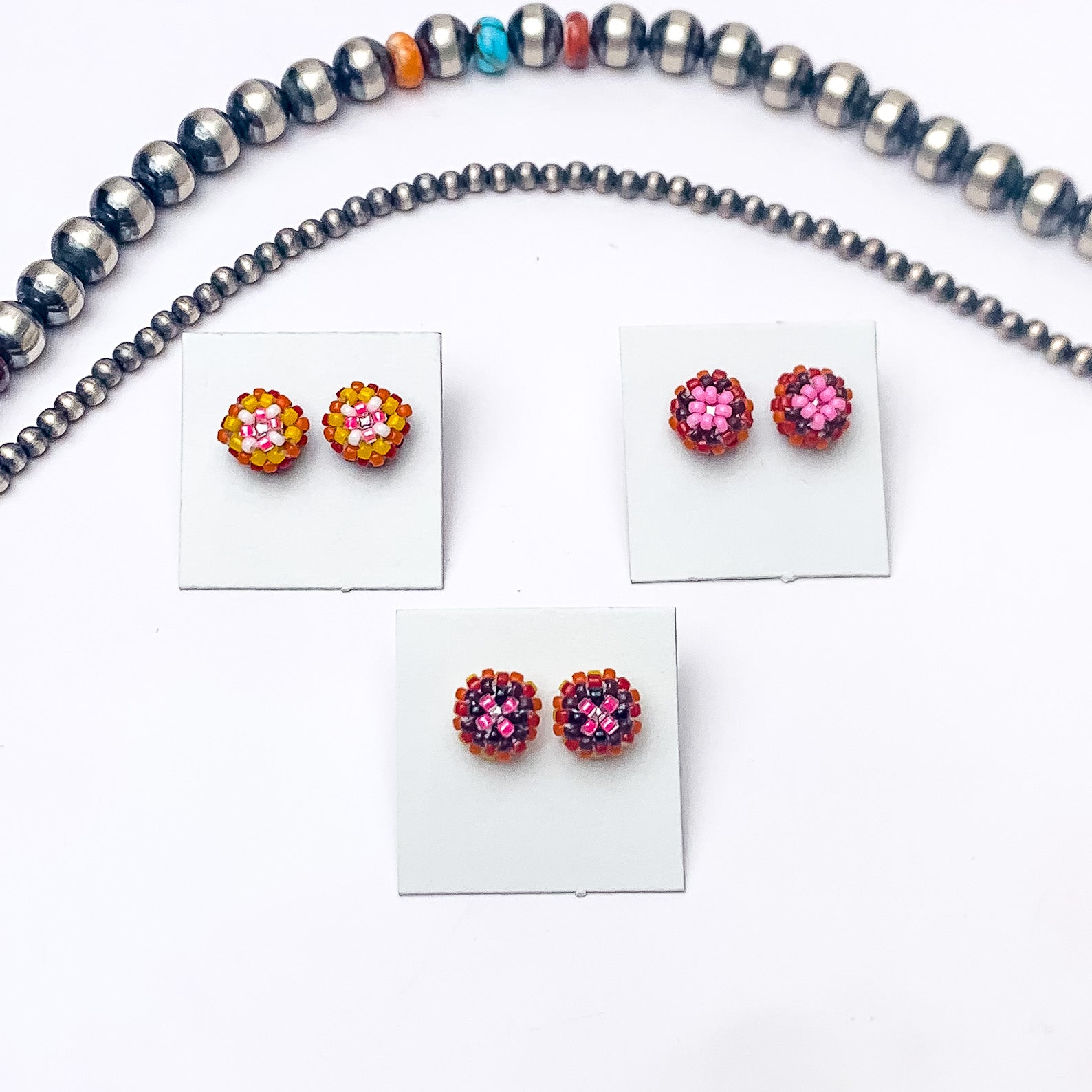 Pictured are four pairs of circle, beaded stud earrings in a mix of orange and pink beads that are different in each pair. These earrings are pictured on a white background with silver beads above the studs.