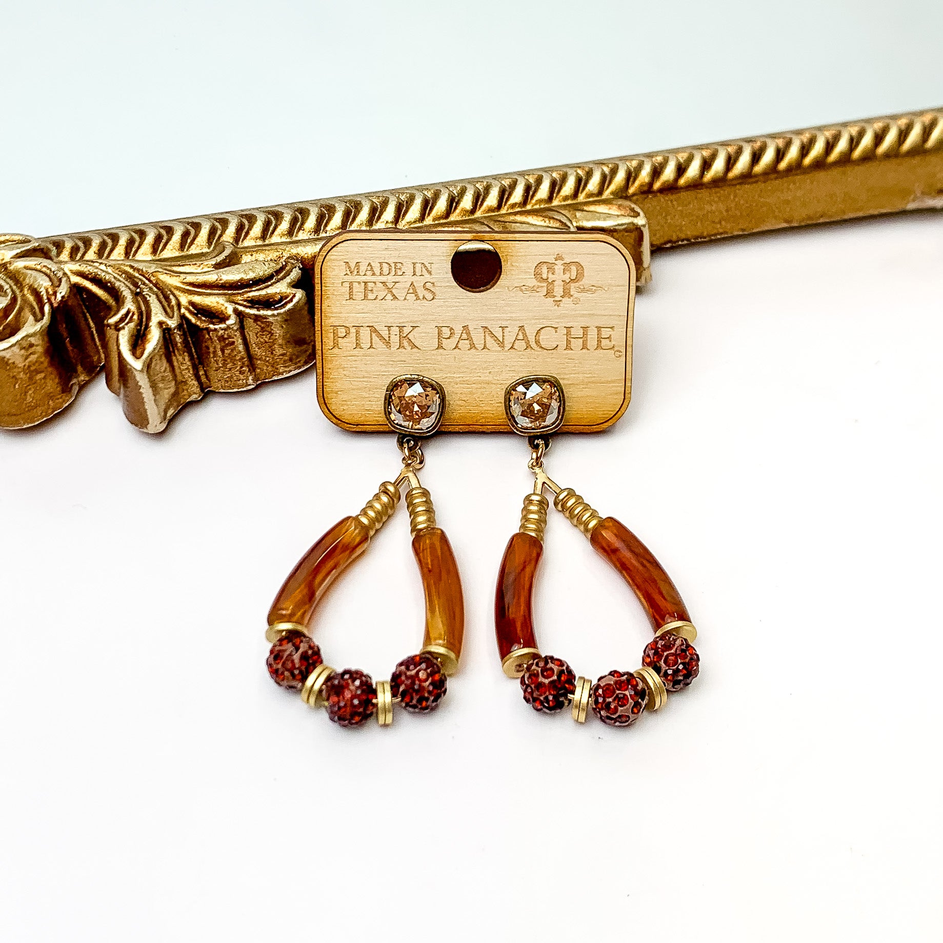 Golden shadow cushion cut crystal post earrings with a teardrop dangle. The teardrop dangle includes gold beads, brown crystal beads, and tortoise bamboo beads. These earrings are pictured on a wooden earring holder on a white background in front of a gold mirror. 