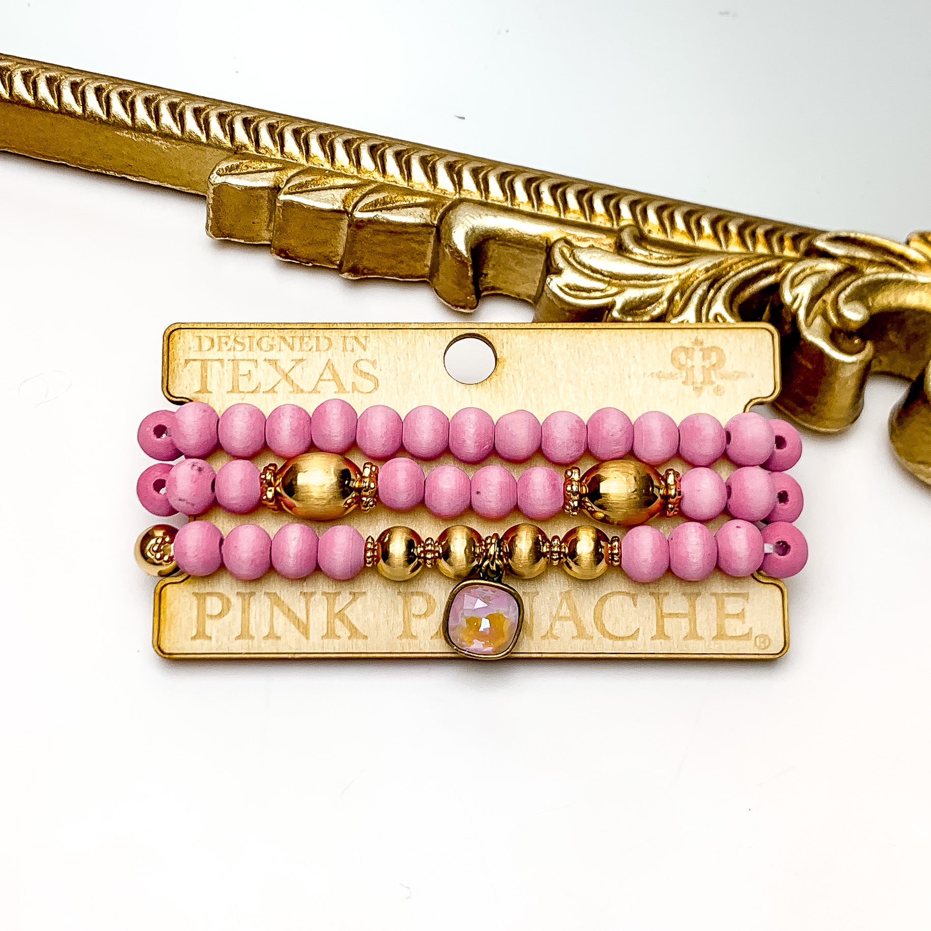Pink Panache Three Strand Pink Wood and Gold Beaded Bracelet With a Dusty Pink Cushion Cut Drop. Pictured on a white background with the top of the bracelets laying against a gold trim frame.