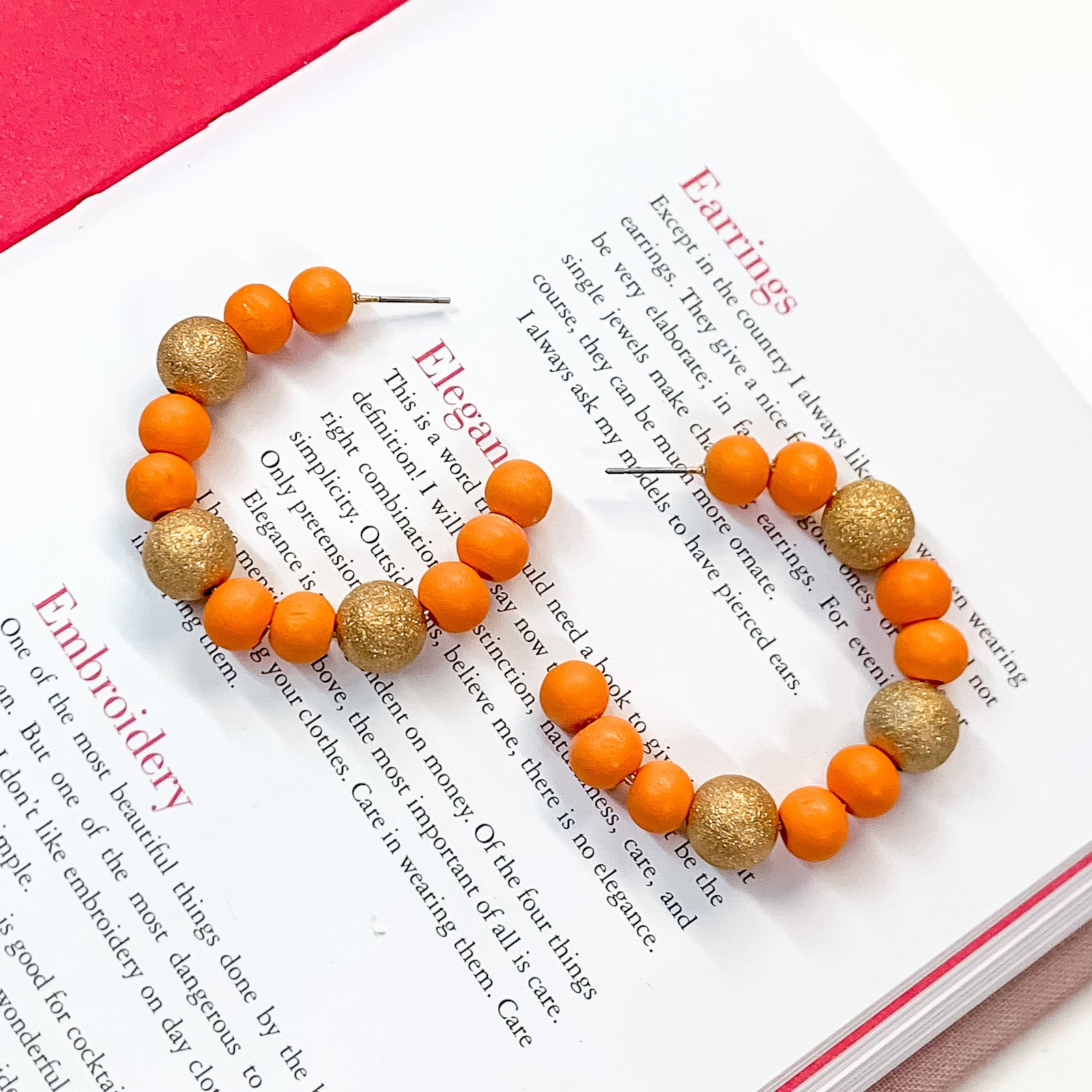 Pictured on an open book is a pair of orange beaded hoop earrings with gold beaded spacers. 