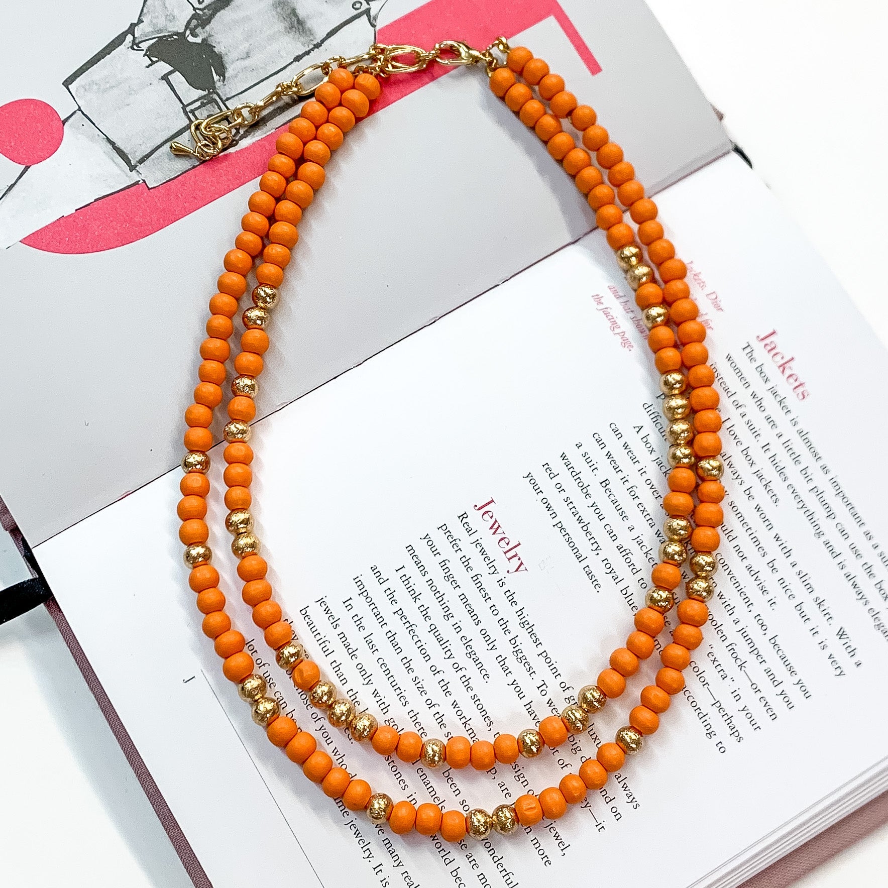 Pictured on an open book is a two strand orange beaded necklace with gold beaded spacers and gold hardware. 