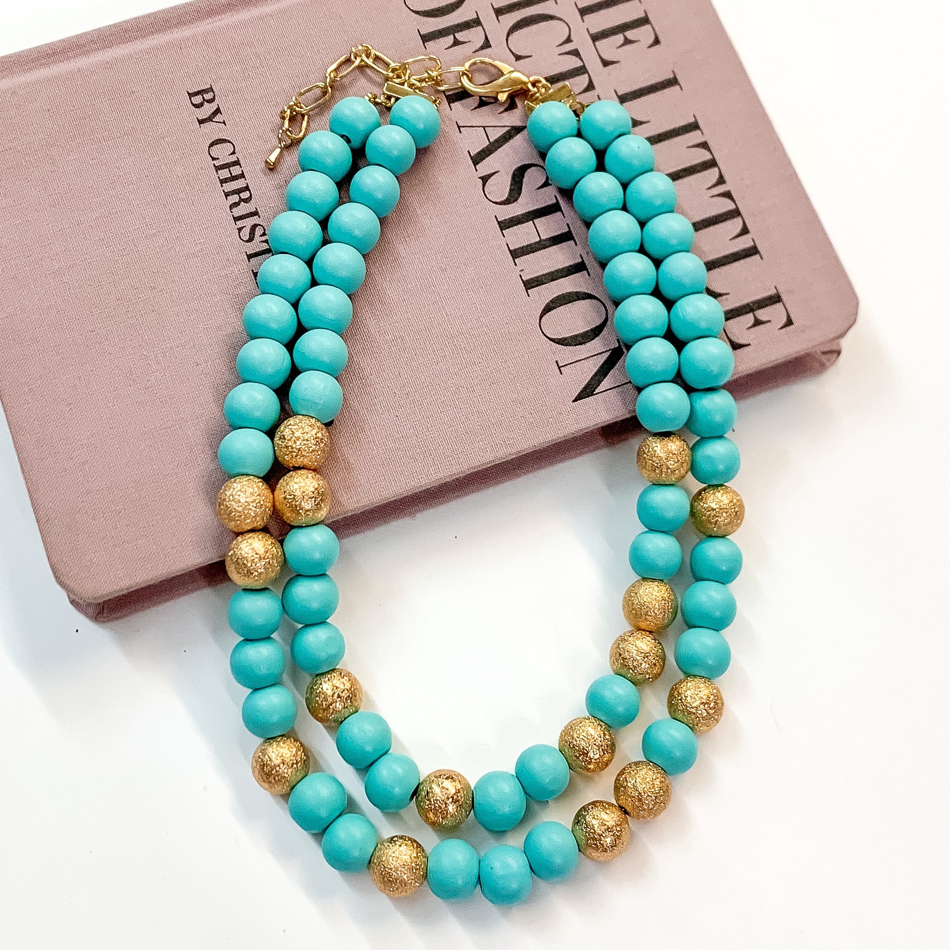 Pictured partially on a mauve colored book on a white background is a two strand sky blue beaded necklace with gold beaded spacers and gold hardware. 