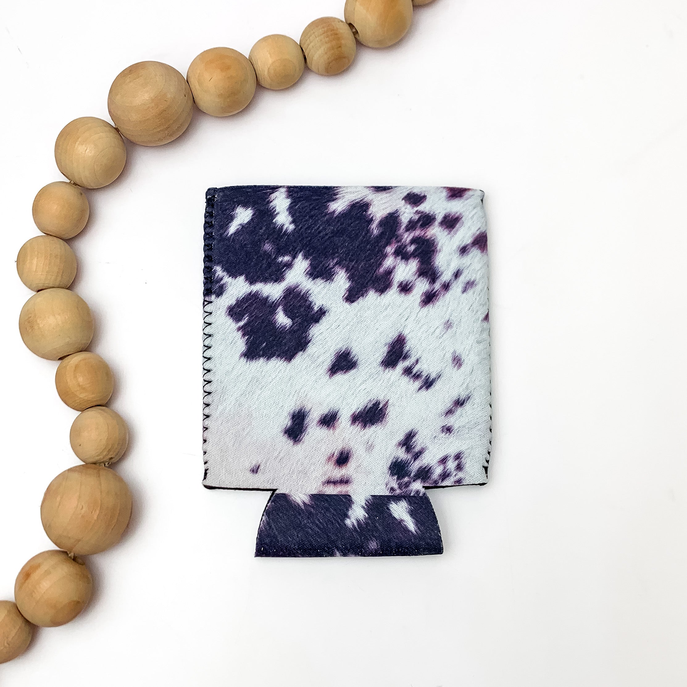 Black and White Cow Print Koozie. Pictured on a white background with wood beads to the left of the koozie for decoration.