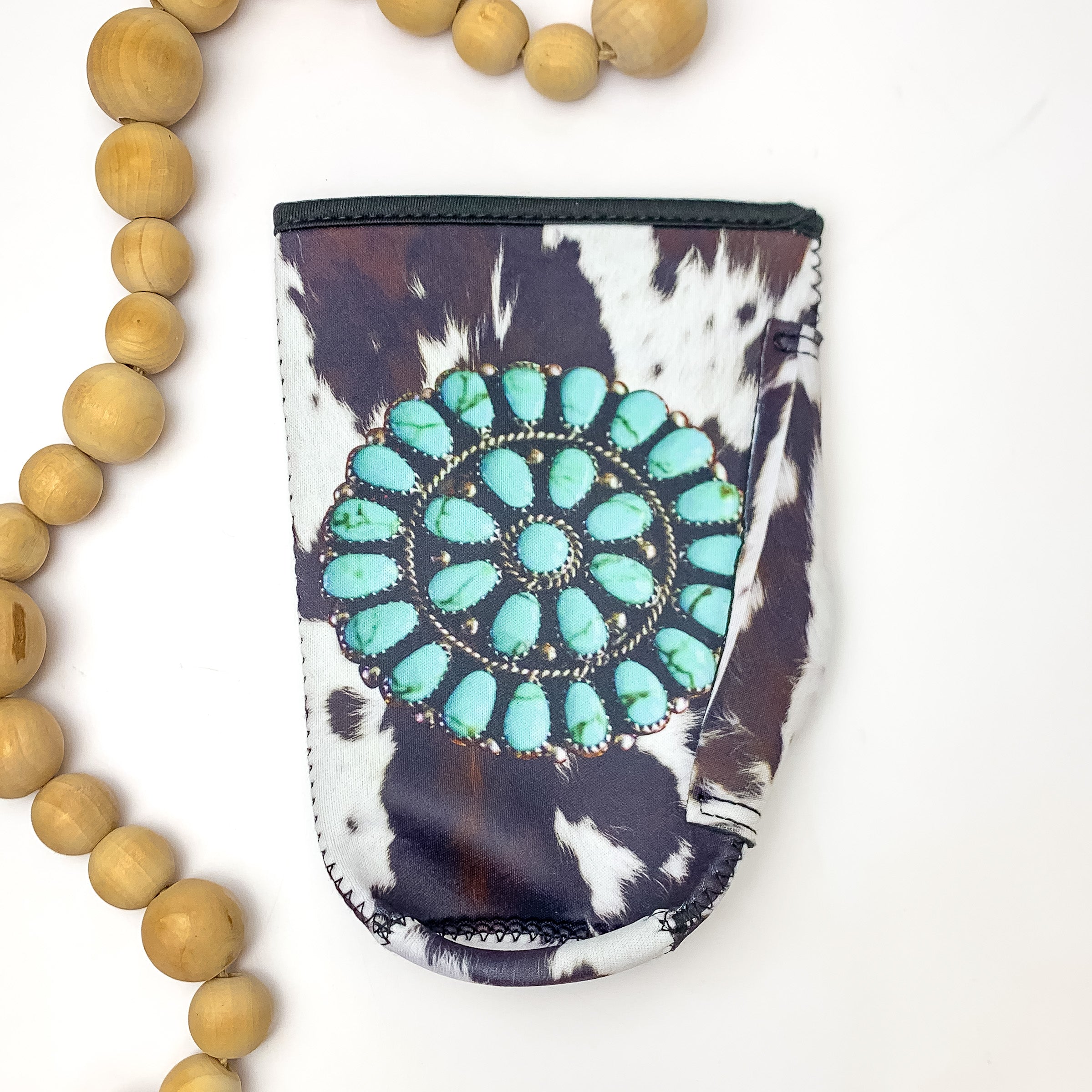Cow Print Tumbler Drink Sleeve With Turquoise Circular Decoration. Pictured on a white background with wood beads on the left for decoration.