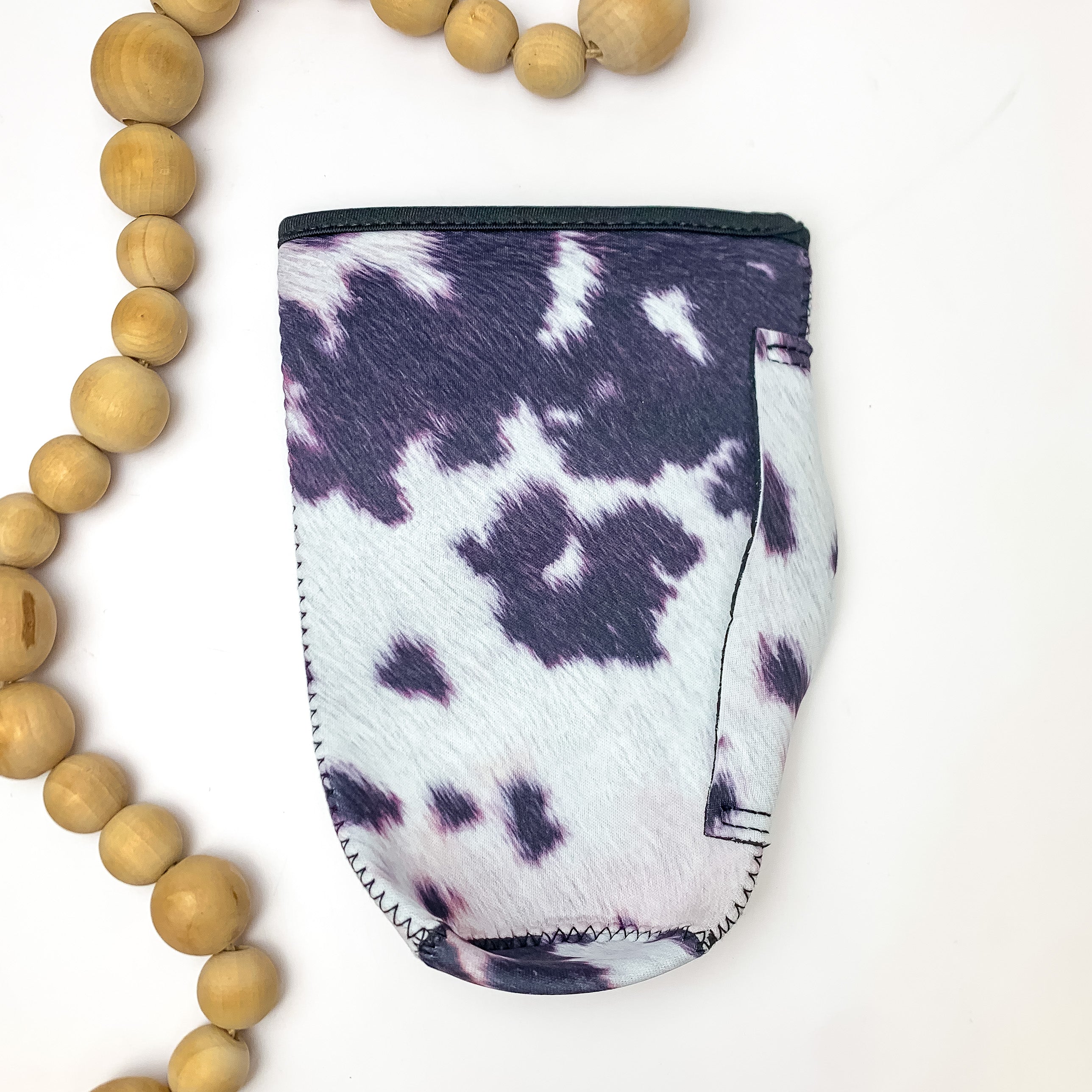 Cow Print Tumbler Drink Sleeve. Pictured on a white background with wood beads next to the koozie for decoration.
