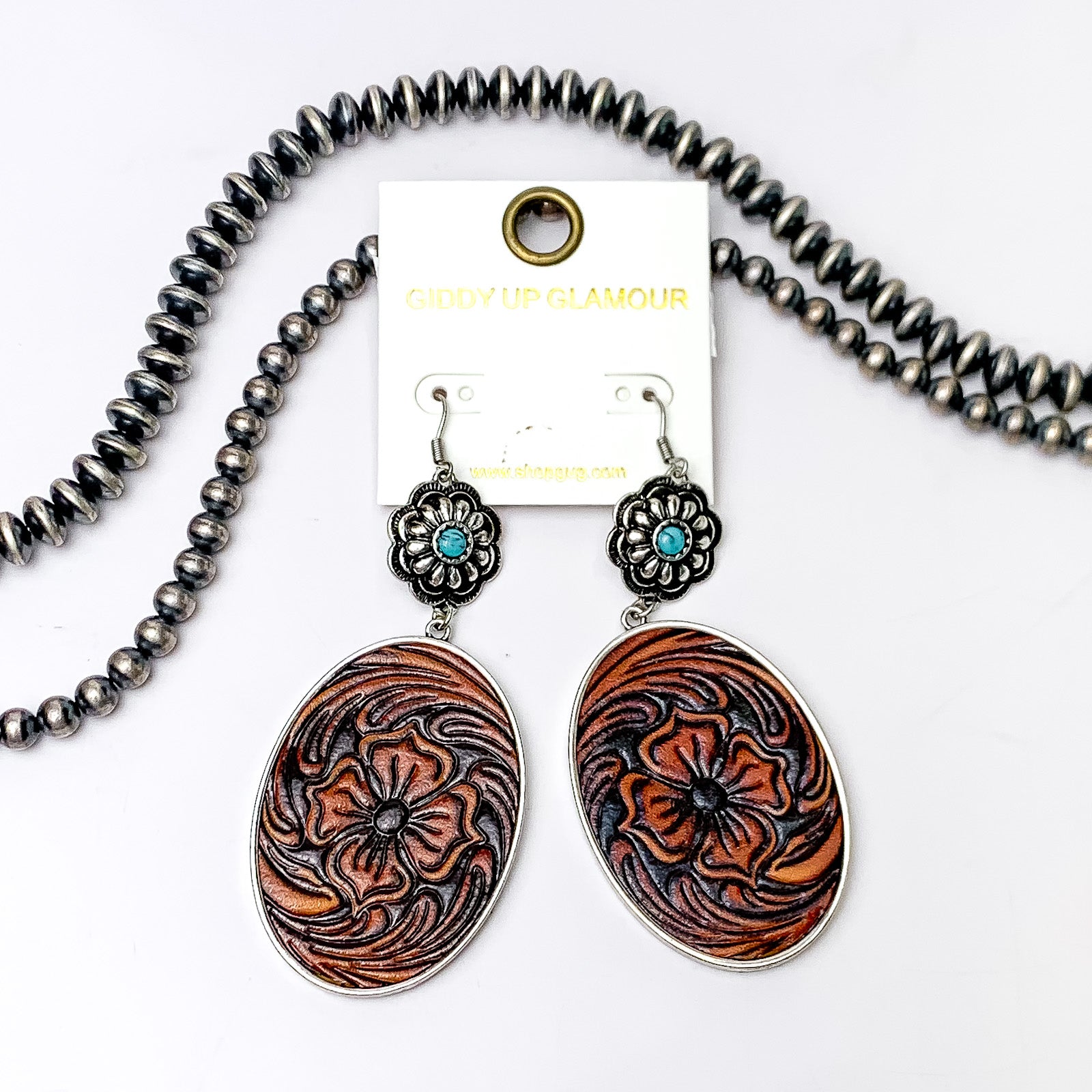 Silver concho dangle earrings with oval pendant. These earrings have a faux leather tooled print. These earrings are pictured on a white background with silver beads at the top of the picture. 
