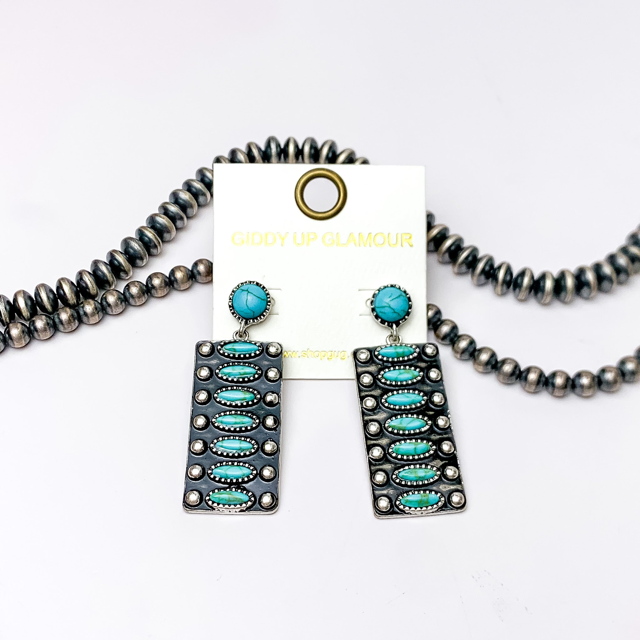 Circle, turquoise post back earrings with a rectangle drop pendant. The rectangle pendant has seven oval turquoise stones. These earrings are pictured on a white background with silver beads at the top of the picture. 