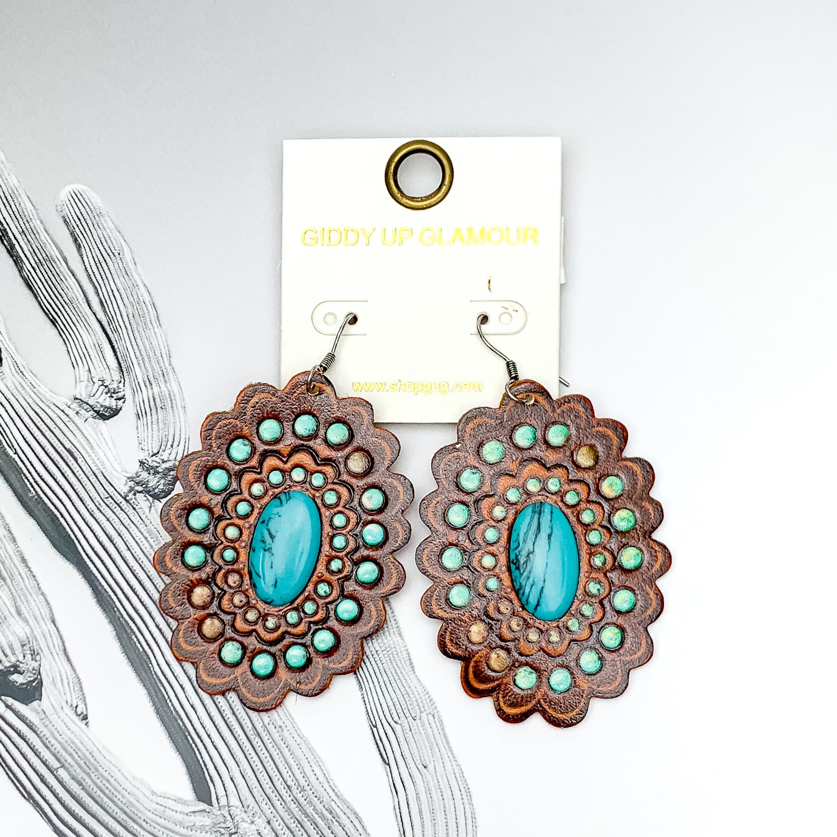Scalloped, oval drop earrings with a silver fish hook. These earrings have a faux leather tooled print with a center turquoise stone. These earrings are pictured on a white and black background. 
