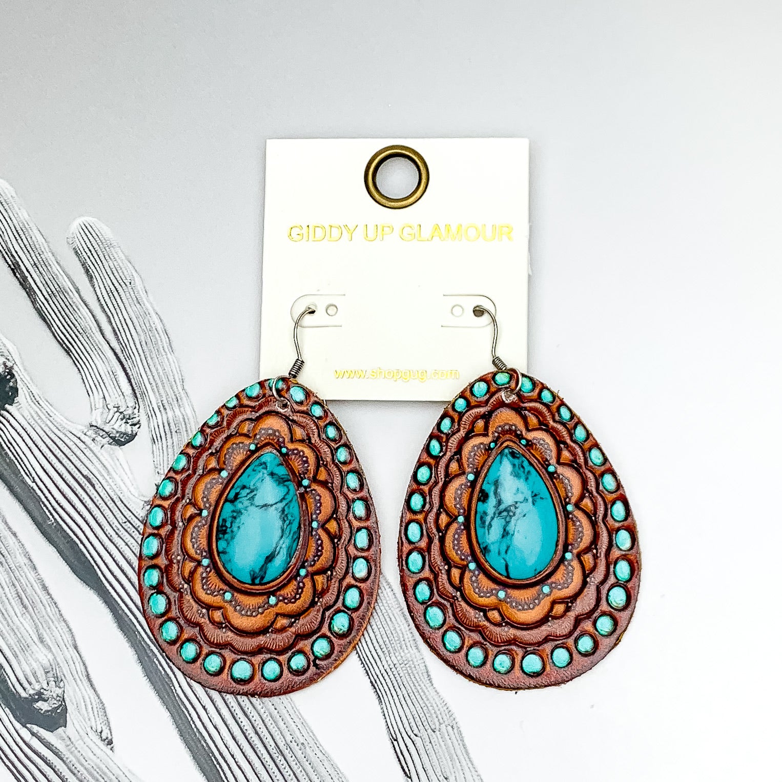 Teardrop earrings with a silver fish hook. These earrings have a faux leather tooled print with a center turquoise stone. These earrings are pictured on a white and black background. 