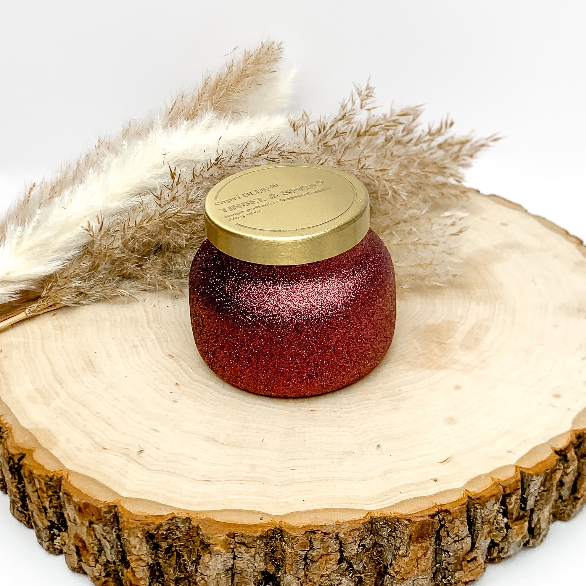 Capri blue 8 oz. jar candle in maroon glitter. The scent of this canfdle is tinsel and spice. The candle is sitting on a wood piece with feathers behind it. The background of the picture is white.