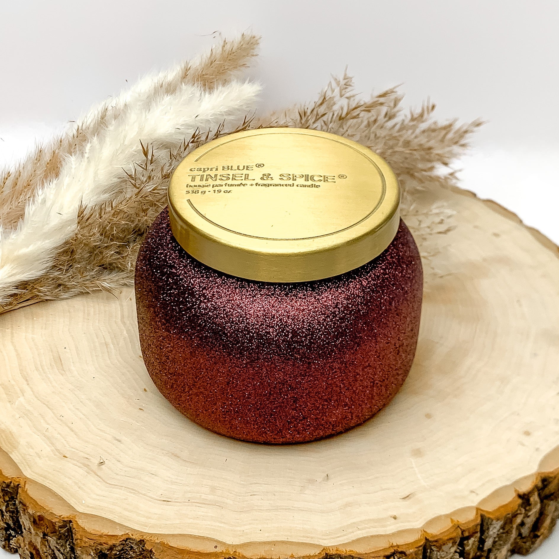 Capri blue 19 oz. jar candle in maroon glitter. The scent of this canfdle is tinsel and spice. The candle is sitting on a wood piece with feathers behind it. The background of the picture is white.