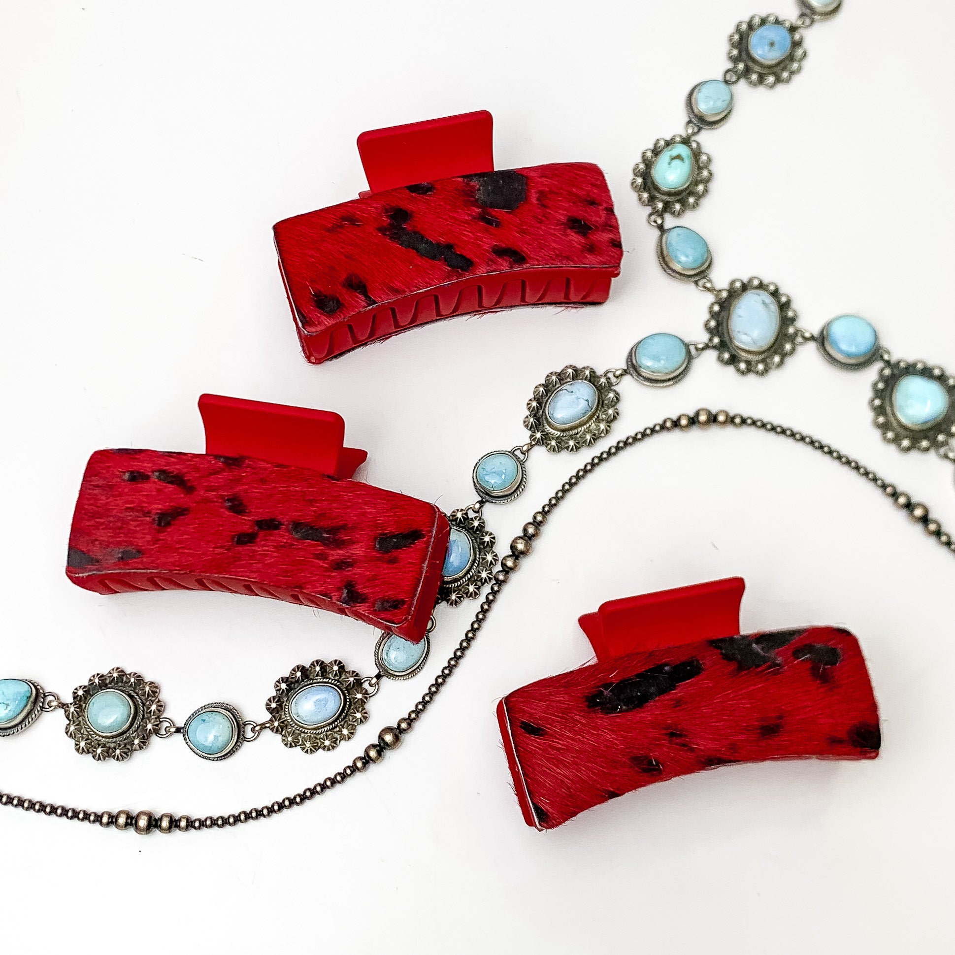 Pictured are three red hair clips with red and black hide patches. These earrings are pictured on a white background with silver necklaces under the clips. 